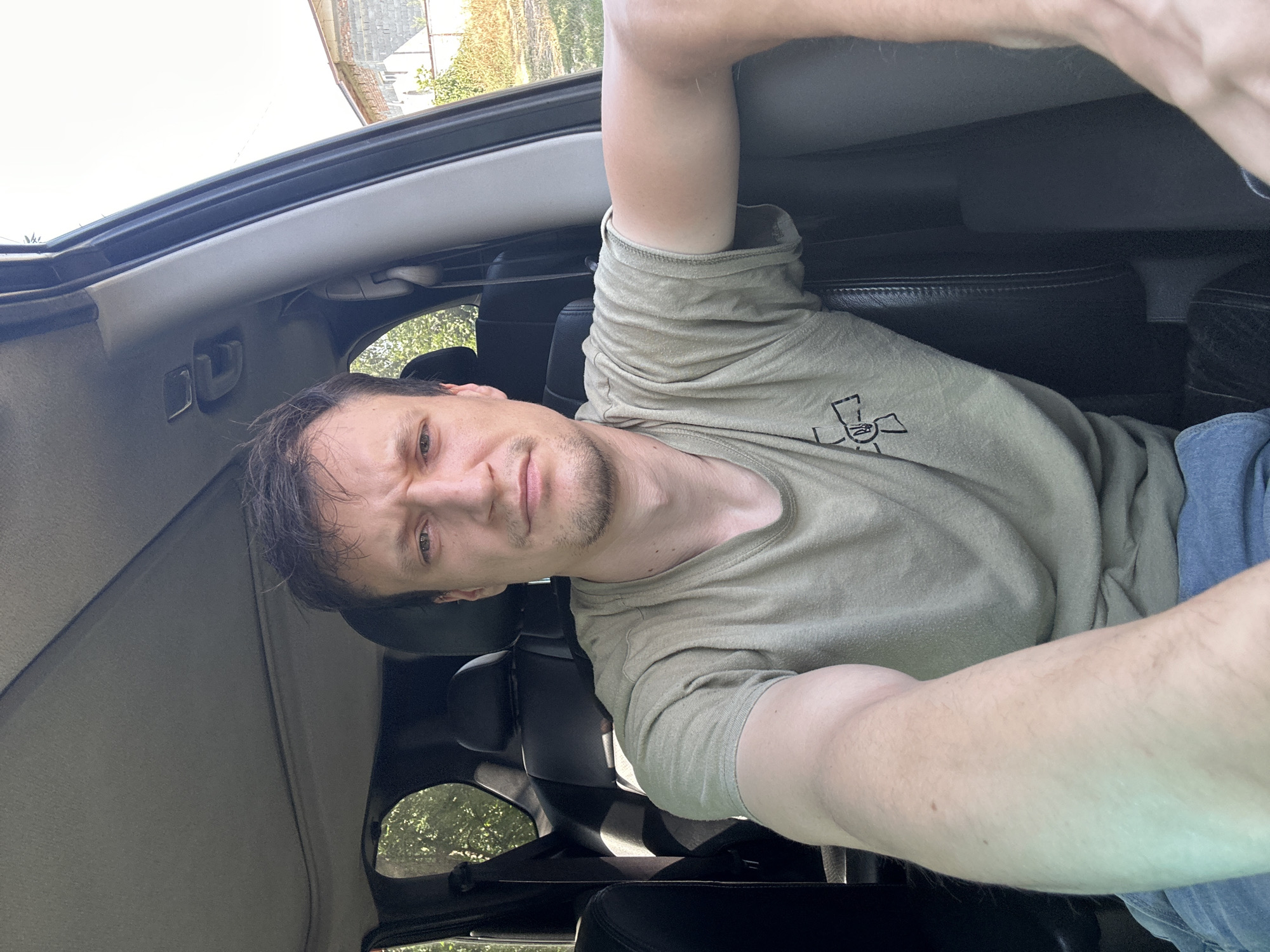 A man is in the driver's seat of a vehicle, looking forward. He has light brown hair and is wearing a tan t-shirt with a black graphic on the left side of his chest. The man's right arm is extended towards the camera. The interior of the vehicle is visible, including black leather seats and part of the sunroof. The exterior of the vehicle shows greenery and a building with a brick wall and a tiled roof. The scene is set during the daytime under a clear sky. The location is Makiv, Ukraine.