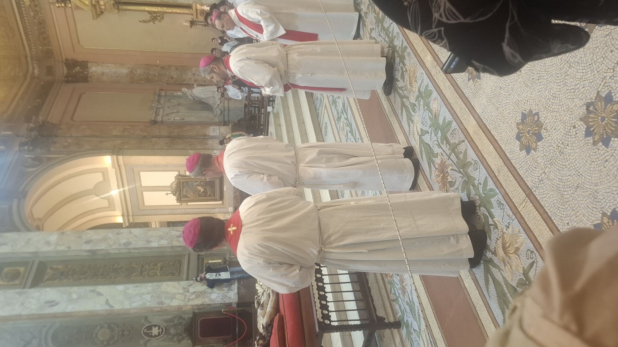 Inside a large building with arches and columns, most likely a cathedral, a group of men, presumably bishops given their red hats and white robes, are standing in a row, most likely bowing or praying. The location is in the city of Buenos Aires, Argentina, although this is not explicitly visible in the image but is based on external knowledge. The floor has a tiled pattern. Red fabric and dark wood furniture are visible on one side. In the background, more people and architectural details are apparent.