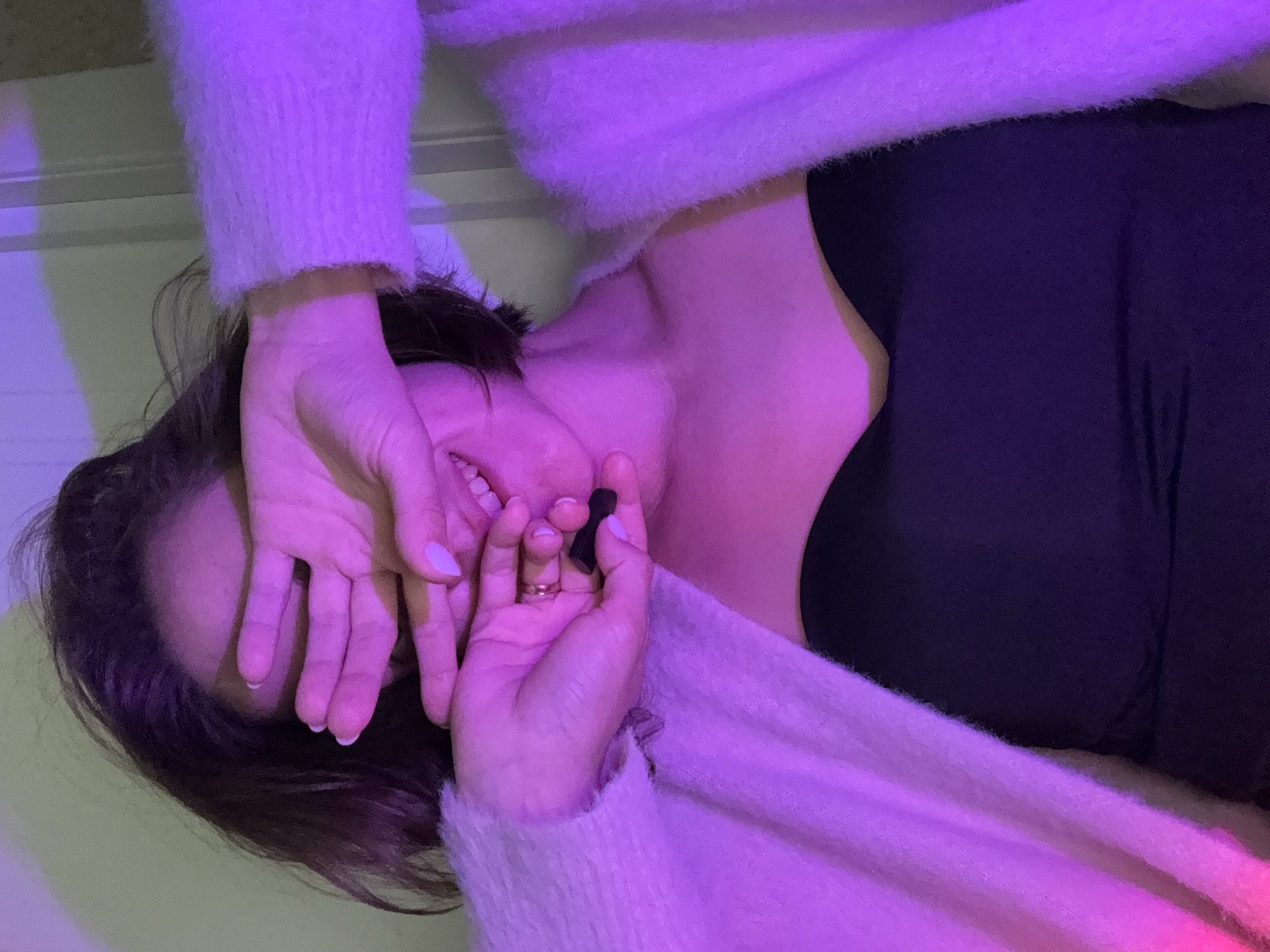 A close-up shot captures a person, likely female, lying down indoors under vibrant purple and pink artificial lighting. The subject's head is tilted, with dark hair framing their face. Their hands are brought up, partially covering their face in a gesture that suggests shyness, amusement, or playfulness, as a faint smile is visible on the exposed part of their mouth. One hand holds a small black object, possibly an earbud, and a gold ring adorns one finger. The nails are painted with a light, possibly white or pale pink, polish.
The person is wearing a light-colored, fluffy, long-sleeved garment, possibly a sweater or cardigan, over a dark top. The purple light casts a strong hue over their skin and clothing. The background is indistinct, appearing to be an indoor wall or surface, with some areas subtly reflecting a greenish undertone through the dominant purple light. The artificial lighting suggests an indoor setting, likely in the evening or at night. No text is visible in the media file. The provided location context is Pervomaysky, Russia, though this is not discernible from the image itself.