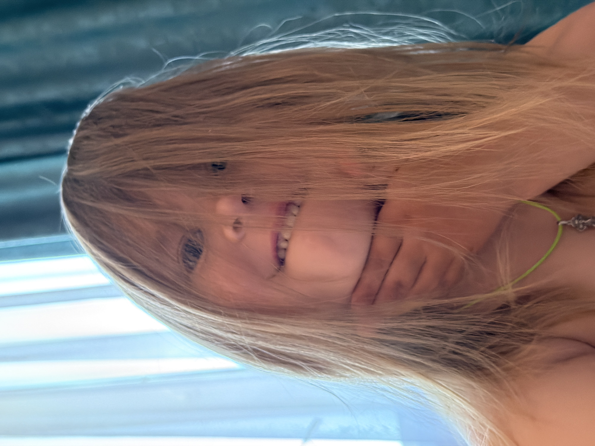 A young child with long, blonde hair obscuring their face is shown in a close-up portrait. Their hair falls forward, covering their eyes and most of their face, with only a glimpse of an eye and a smile visible. The child is wearing a thin, green necklace with a small, silver pendant. Their left hand is raised to their chin, partially supporting their head. The background consists of light blue vertical blinds on the left and a textured teal curtain on the right. The lighting suggests an indoor setting, possibly a home in Chaikovsky, Russia, with natural light filtering through the blinds.