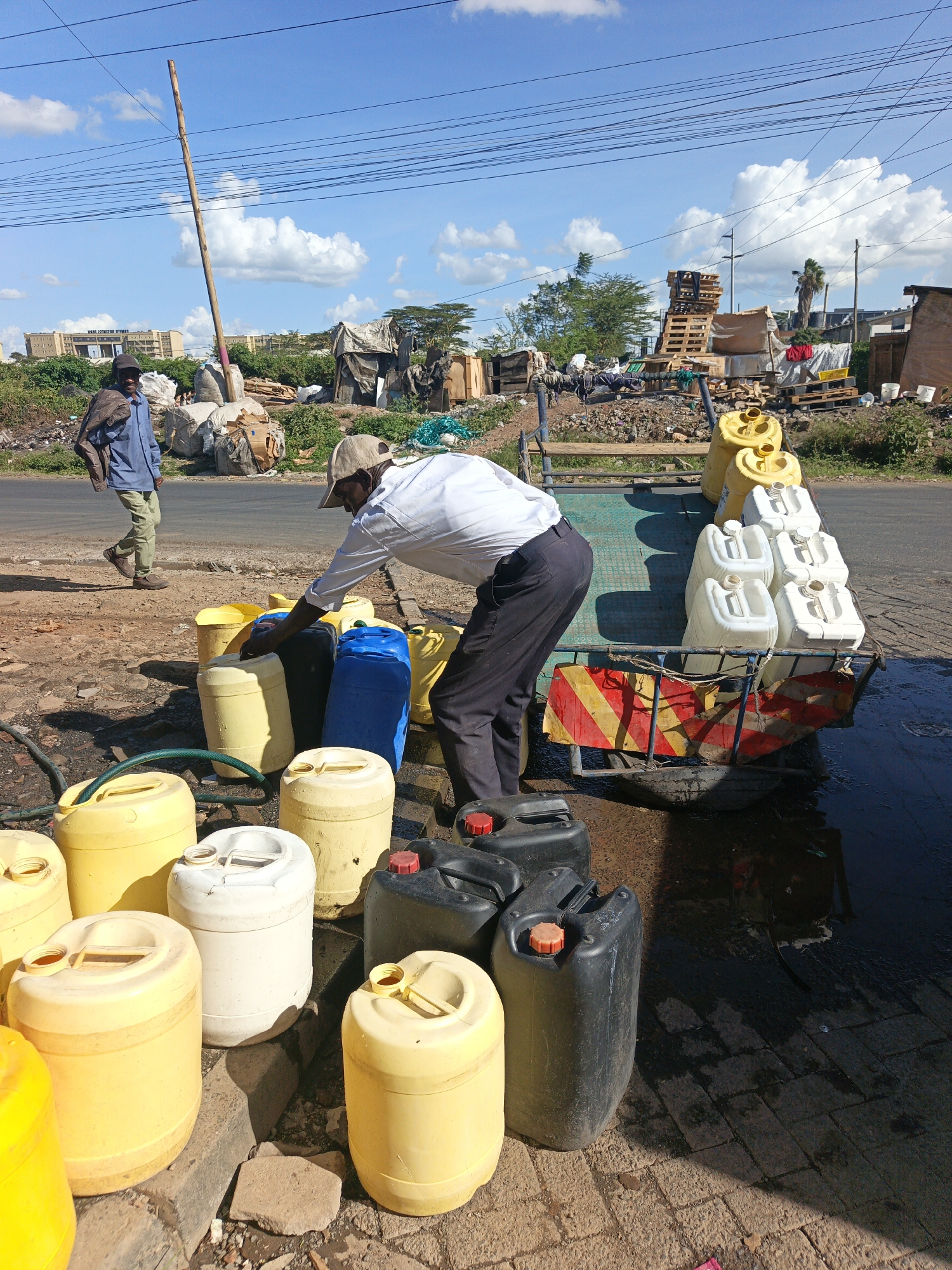 The image shows a man filling yellow, white, blue, and black plastic jerrycans with water from a hose in an urban setting. He is wearing a white shirt, dark pants, and a light-colored cap. Another man walks past in the background, wearing a blue shirt, light pants, and a dark cap, with a jacket slung over his shoulder. The jerrycans are arranged both on the ground and on a small trailer with a reflective red and yellow warning panel, which is hitched to a vehicle, partly visible on the right. Puddles of water are on the ground. Debris, including wooden pallets and various materials, is scattered in the background, along with buildings, suggesting a slum or informal settlement. A tall wooden pole and power lines are overhead. The scene is located on a paved area next to a road, potentially within the city of Nairobi, Kenya, given the general context of water distribution and infrastructure.