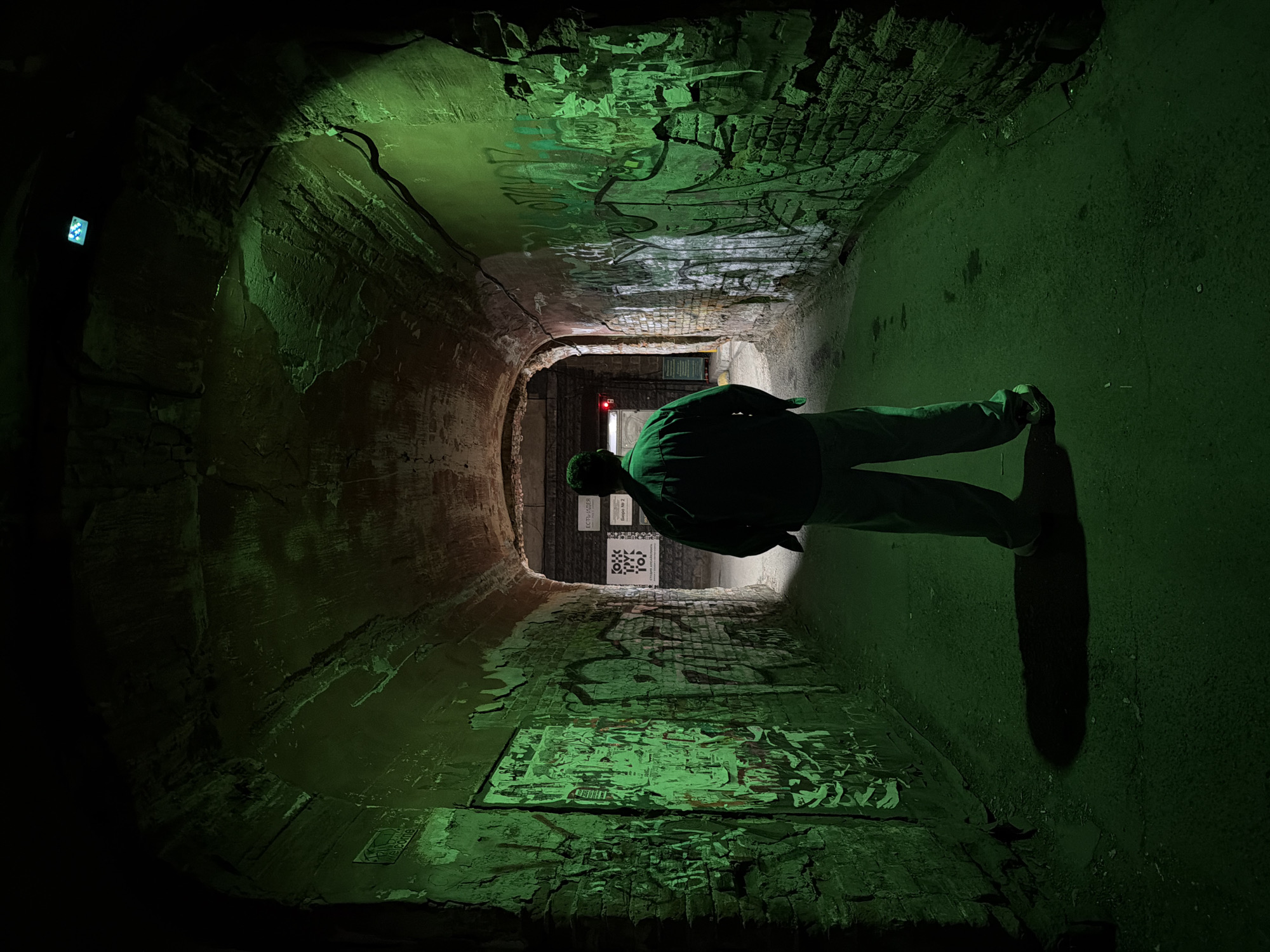 A person stands at the entrance to a dark, arched passageway in Krasnoyarsk, Russia. The passageway has rough brick walls, adorned with graffiti and posters. The area is illuminated with green light, emanating from above and casting long shadows. At the far end, signs are visible on the back wall. A red light glows above the exit.
