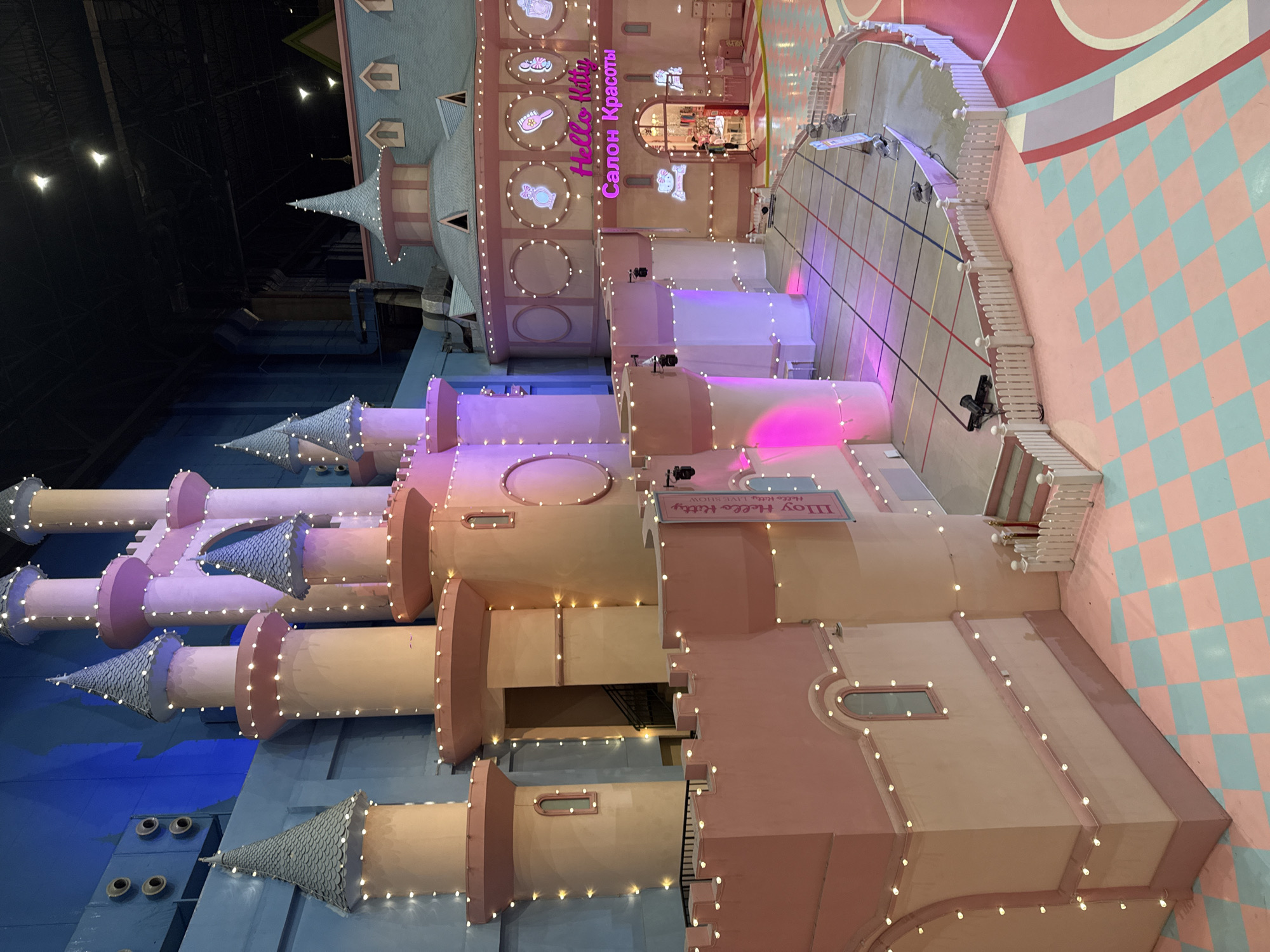 The image captures a colorful and whimsical scene within the "Island of Dreams" indoor amusement park in Moscow, Russia. The main subject is a large, ornate structure resembling a castle or series of interconnected buildings, adorned with pastel colors (pink, peach, blue) and decorative elements like turrets, rounded roofs, and string lights. The architecture suggests a theme park or entertainment venue designed for children or families.
The buildings feature signs in Russian script, one reading "Салон Красоты" (Salon Krasoty), which translates to "Beauty Salon," and another sign that says "Шоу Hello Kitty". The structure appears to house shops or attractions related to Hello Kitty.
The surrounding environment includes a checkered pink-and-blue floor pattern, a curved area with lines painted on the ground, potentially for queues or crowd control, and what appears to be a stage or elevated area. There are no people visible in the image.