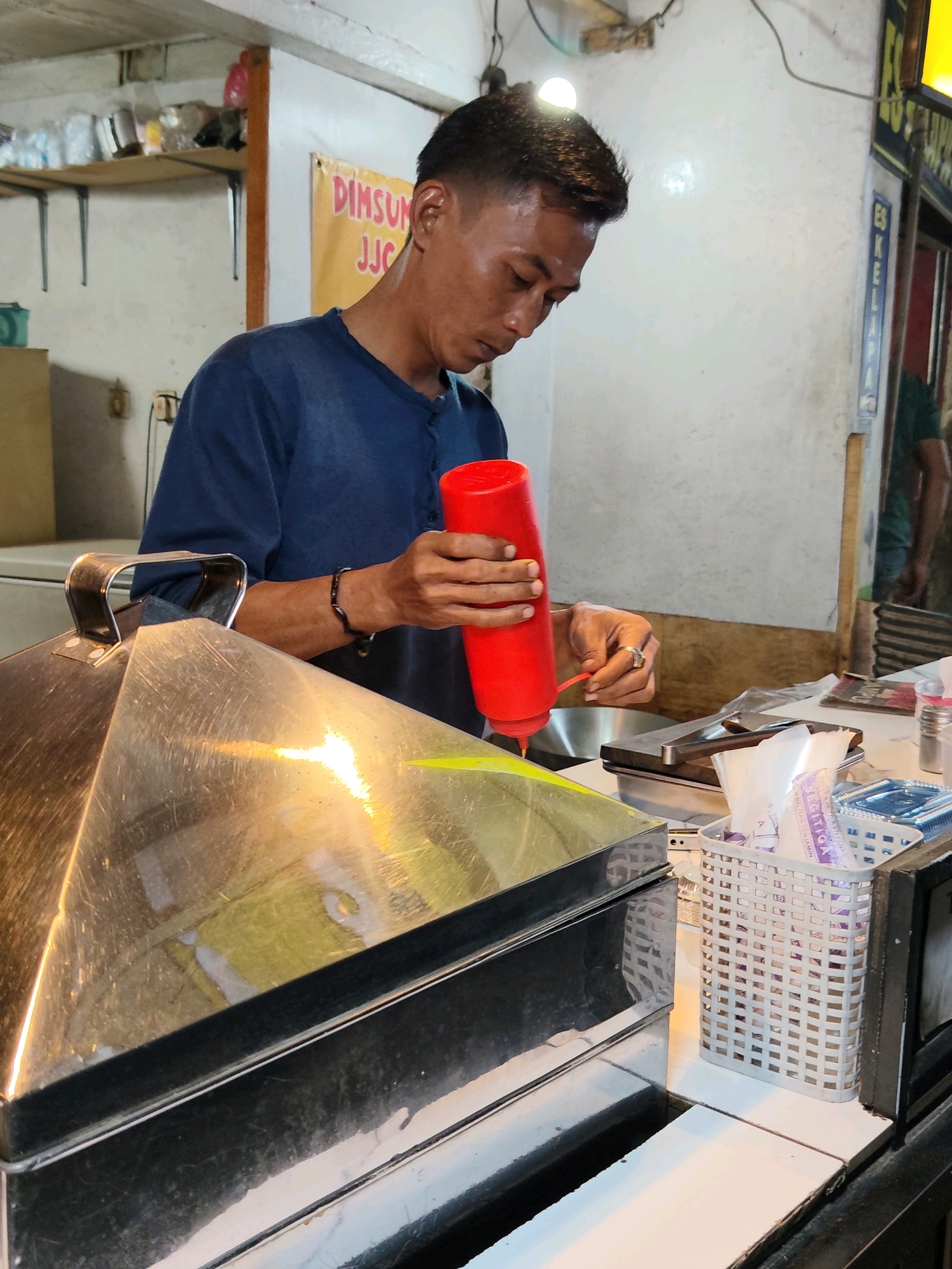 A man, possibly a street food vendor, is seen in a dimly lit, enclosed space, likely a food stall or small eatery, in the city of Purwakarta, Indonesia. He is wearing a navy blue, long-sleeved shirt and is focused on dispensing a red sauce from a squeeze bottle into a metal container or dish. His left hand holds the bottle, while his right hand is positioned near the nozzle, suggesting he is controlling the flow of the sauce. A silver, geometric-shaped lid of a food steamer or warmer is prominently in the foreground, reflecting the overhead lighting. Behind him, a yellow banner with red lettering that reads "DIMSUN JJC" is visible on a white wall, indicating the name of the establishment or product. The environment appears utilitarian, with shelves in the background holding various items, and a metal railing or counter edge. The overall scene suggests a preparation or serving process for food, possibly dim sum given the banner.