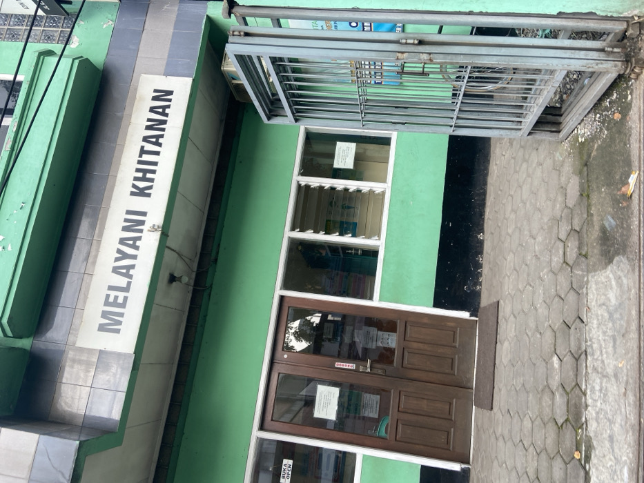 The image shows the exterior of a building in Kecamatan Cimahi Selatan, Indonesia (latitude: -6.932, longitude: 107.564). The building is painted a light green color and has a sign above the entrance that reads "MELAYANI KHITANAN." The sign is white with black lettering. There is a double wooden door with a sign that says "BUKA OPEN" located on the left side of the entrance. The entrance is flanked by two windows that are open. The windows have white vertical louvers. A metal gate is on the right side of the entrance. The ground is covered in gray hexagonal pavers. There is a small brown welcome mat located in front of the entrance. The building appears to be a medical clinic or a health facility. There are no people visible in the image. The scene is calm and devoid of any activity. There is no other information available about the scene.