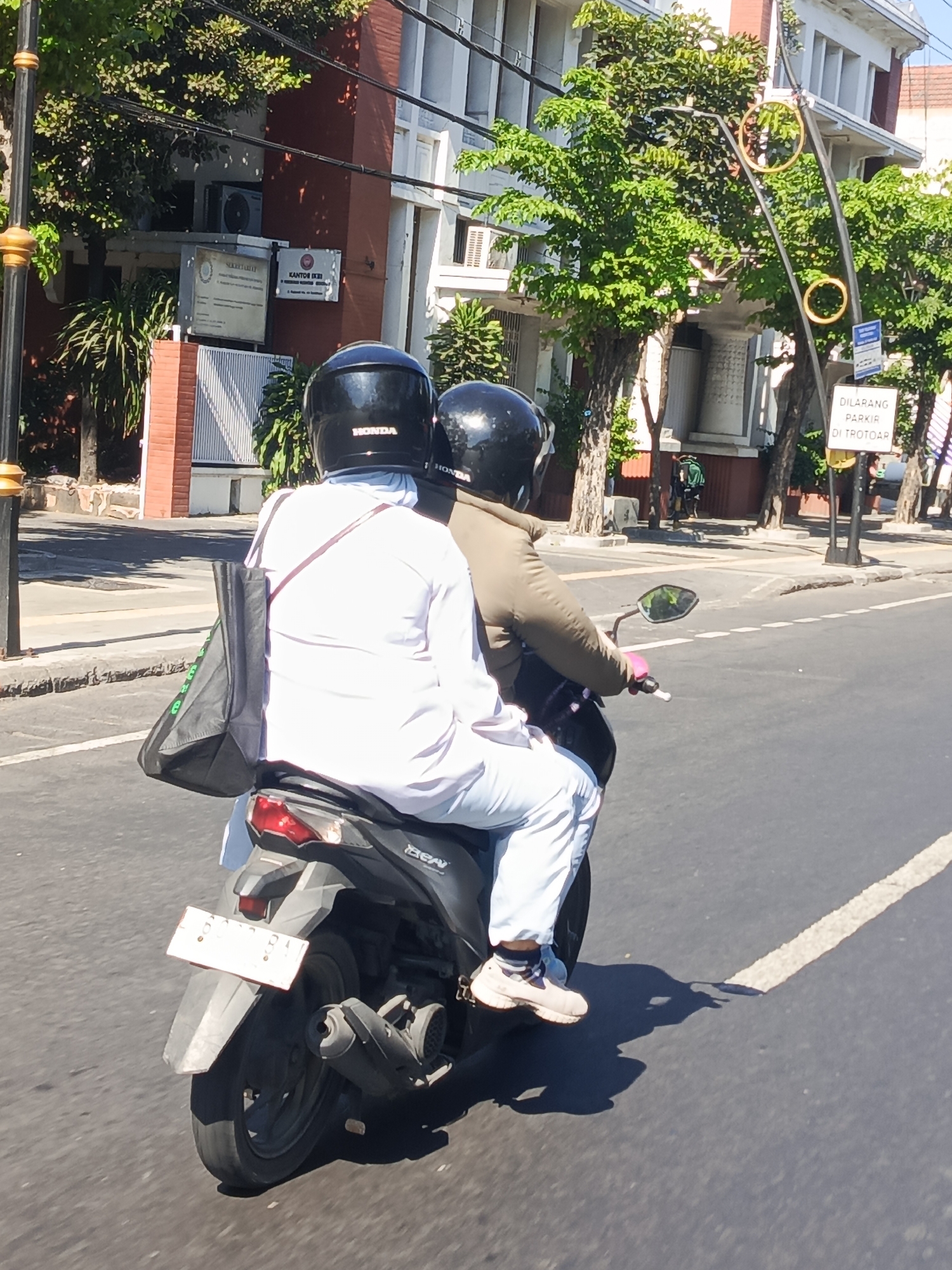 Two individuals are riding a black Honda Beat scooter on a city street in Surabaya, Indonesia, during daylight. The person in the front is wearing a white long-sleeved shirt, light blue pants, and white sneakers, with a black helmet. They are carrying a black tote bag over their left shoulder. The person in the back is wearing a brown jacket and a black helmet, with their hands on the driver's shoulders. The scooter has a white license plate. The street is paved with asphalt and has a white lane marking. In the background, there is a building with red brick and white accents, and lush green trees. A sign with Indonesian text that translates to "PARKING IS FORBIDDEN ON THE SIDEWALK" is visible on a pole. The sun is casting shadows on the road, indicating a clear, sunny day. The overall scene depicts a typical urban commute in Surabaya.