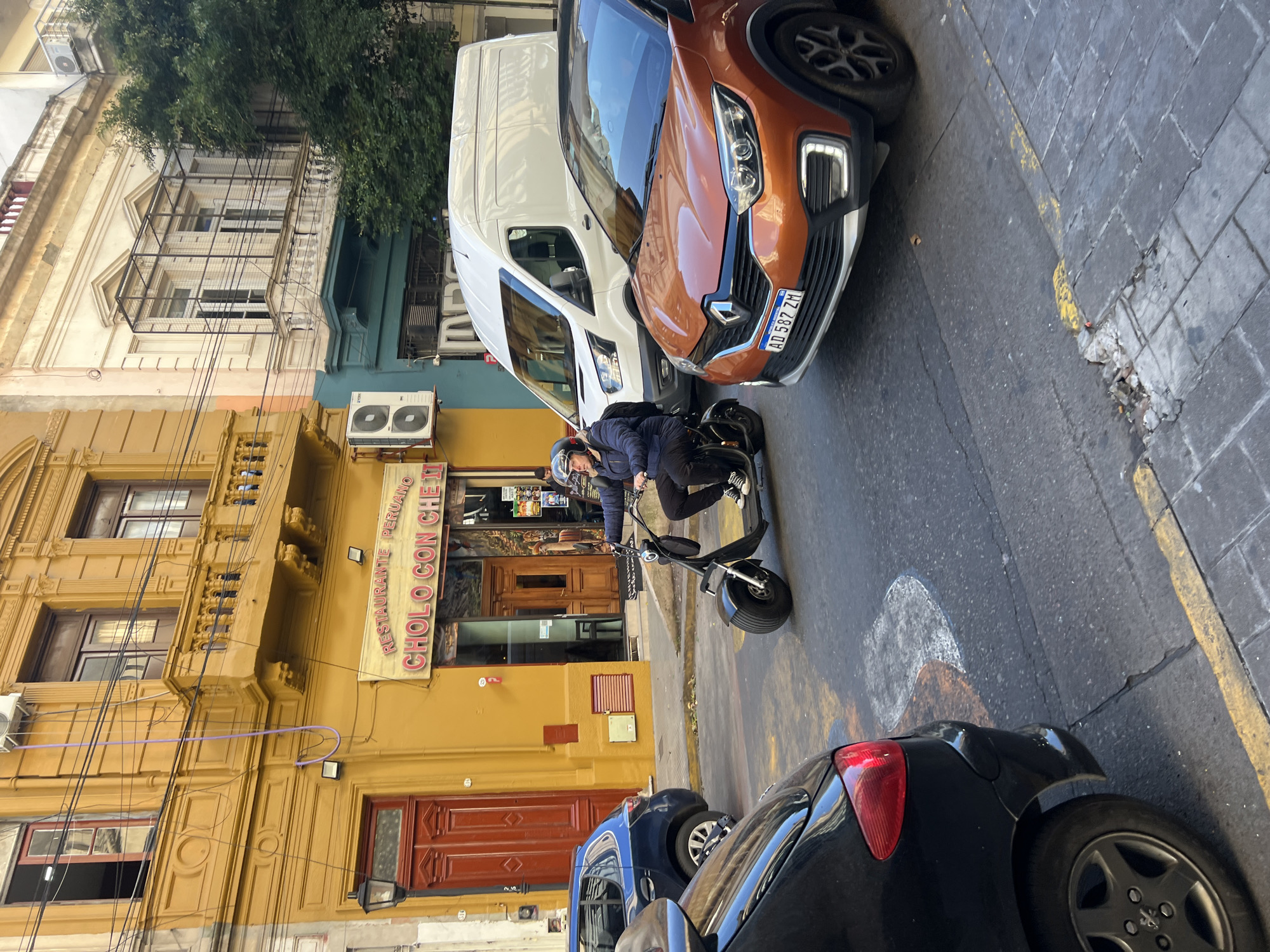 The image shows a street scene in Buenos Aires, Argentina. A person wearing a helmet and dark clothing is riding a black scooter on the street. Cars are parked on both sides of the street, including a white van and a brown Renault with a license plate "AD 587 ZM."
Behind the scooter rider is a yellow building with ornate architectural details. A sign for "RESTAURANTE PERUANO CHOLO CON CHE III" is visible above a dark wood door. The street surface is paved with rectangular stones or pavers, showing signs of wear and patched areas. Overhead, power lines crisscross in front of the buildings.