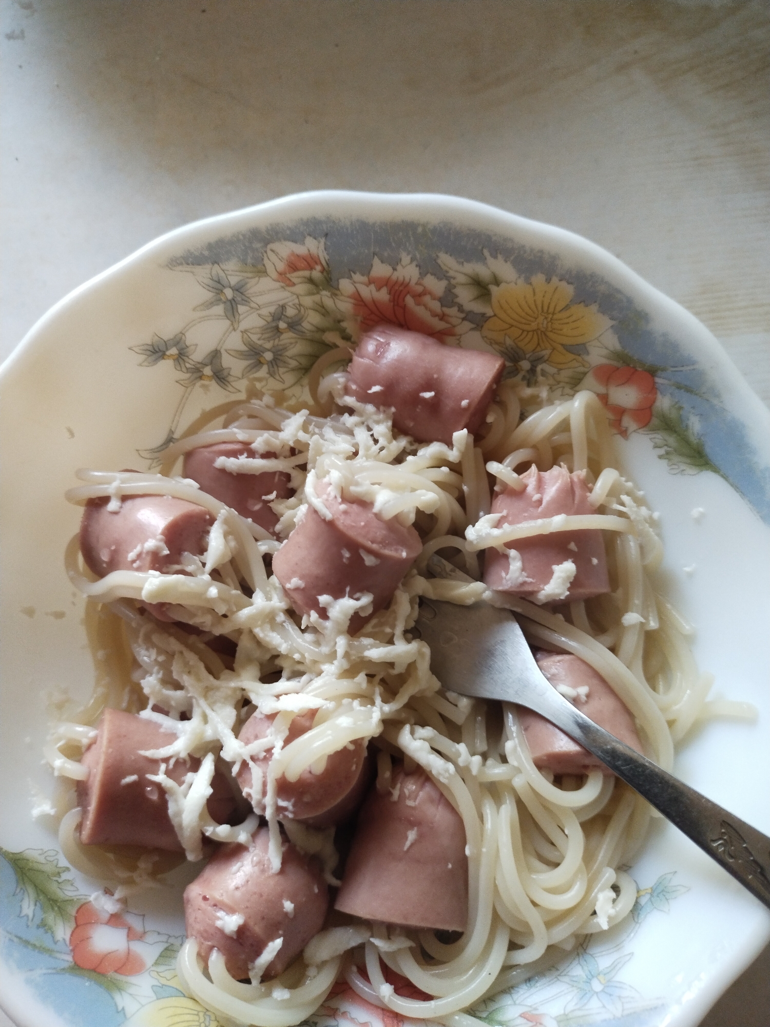 This image depicts a close-up, overhead view of a meal served in a decorative white ceramic bowl, located in the city of Horishni Plavni, Ukraine. The bowl contains light-colored spaghetti or thin noodles mixed with several thick, cut pieces of processed sausage, likely frankfurters or hot dogs, which are pinkish-brown in hue. A substantial amount of white, finely shredded or grated substance, consistent with cheese, is liberally scattered over the pasta and sausage. A silver-colored metal fork is inserted into the food, positioned diagonally across the bowl with its tines submerged. The rim of the bowl features a floral pattern with blue, orange, yellow, and green elements against a muted blue-grey band. The meal rests on a light-colored, subtly textured surface, possibly a wooden tabletop or countertop. The scene is brightly lit, with soft shadows.