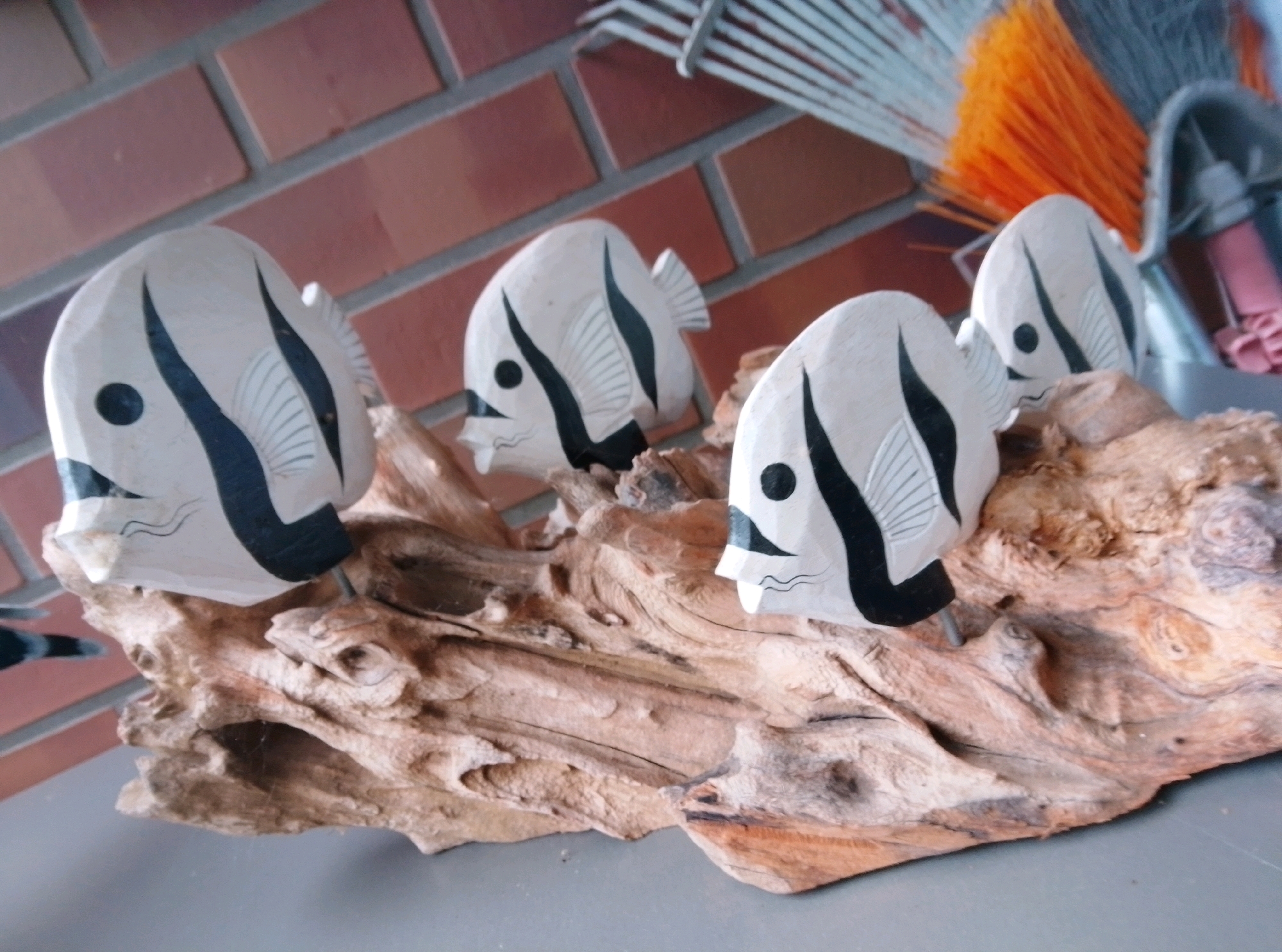 The image shows a close-up, slightly angled view of four wooden fish sculptures placed on a piece of driftwood. The fish are predominantly white with black stripes and details, resembling butterflyfish. Three of the fish are in focus, arranged in a row on the driftwood, with the fourth fish slightly out of focus in the background. The driftwood itself is rough and textured, with various cracks and natural formations. Behind the driftwood and fish, a brick wall is visible, appearing to be red or reddish-brown. To the upper right, part of a brush with orange bristles and a metal handle is visible, suggesting the scene might be indoors or in a covered outdoor area. The lighting is soft and appears to be from the side, casting subtle shadows. The overall setting does not provide specific indicators of a location within the city of Wedel, Germany, as there are no identifiable landmarks or context clues related to the city.