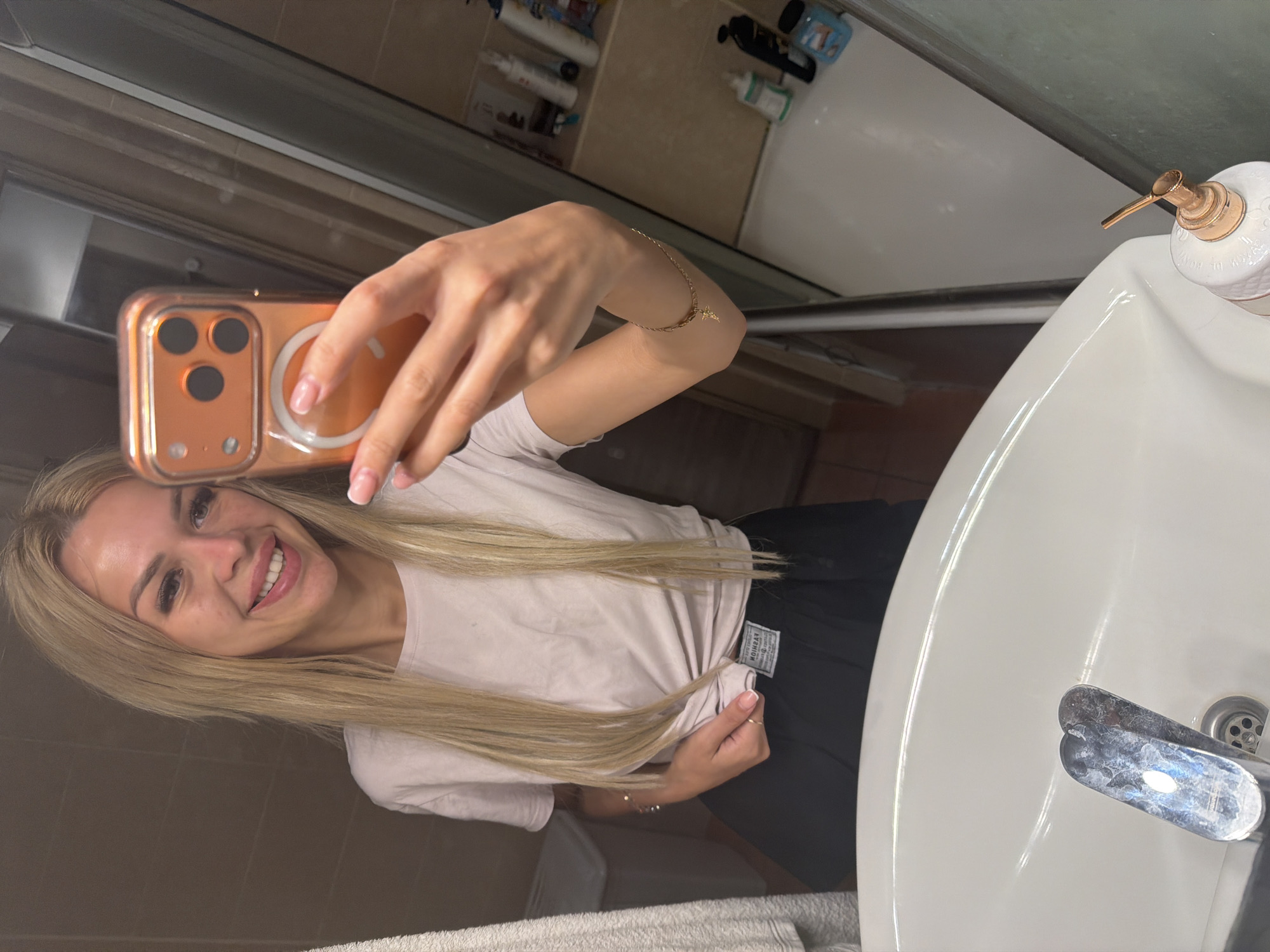A young woman with long blonde hair takes a selfie in a bathroom, holding an orange phone in her right hand. She is wearing a light pink t-shirt and black shorts. Her left arm is bent, and she has a thin gold bracelet on her wrist. She is smiling, with her teeth showing. The background shows a tiled wall, a white bathtub, and a sink with a soap dispenser. The scene appears to be indoors, likely in a home bathroom, possibly in Kyiv, Ukraine. The lighting suggests it is daytime, but no external weather cues are visible. The overall mood is casual and happy. There is a label on the woman's t-shirt that is partially obscured and unreadable.