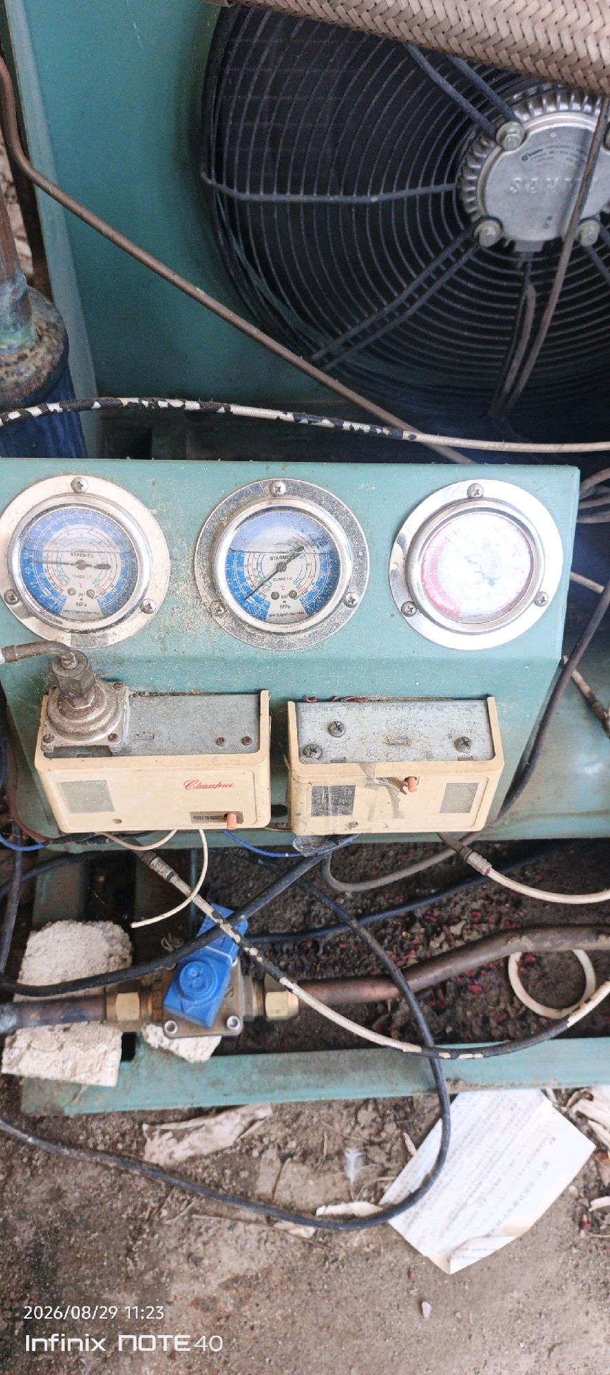This is a medium shot, eye-level, outdoor photograph taken in Patampanua, Indonesia. The image focuses on a piece of industrial machinery, likely an air conditioning unit, given the presence of gauges, copper piping, and a large fan at the top. The machinery is teal green and weathered, with visible rust and dirt. Three pressure gauges are mounted on the front panel. The leftmost gauge appears to be reading around 30 psi, and the middle gauge around 40 psi, both with blue colored scales indicating refrigerant pressure. The rightmost gauge has a red and white scale and is difficult to read clearly due to glare, but it also appears to be a pressure gauge. Below the gauges are two beige electrical boxes with visible wiring. One box has a label that reads "Chuanfai" and a small orange button. A blue valve with a black handle is visible in the lower portion of the image, connected to copper pipes. The ground beneath the machinery is rough and appears to be dirt mixed with small debris. A piece of white paper with text, possibly instructions or a label, lies on the ground to the right. The time stamp on the image indicates 2026/08/29 11:23.