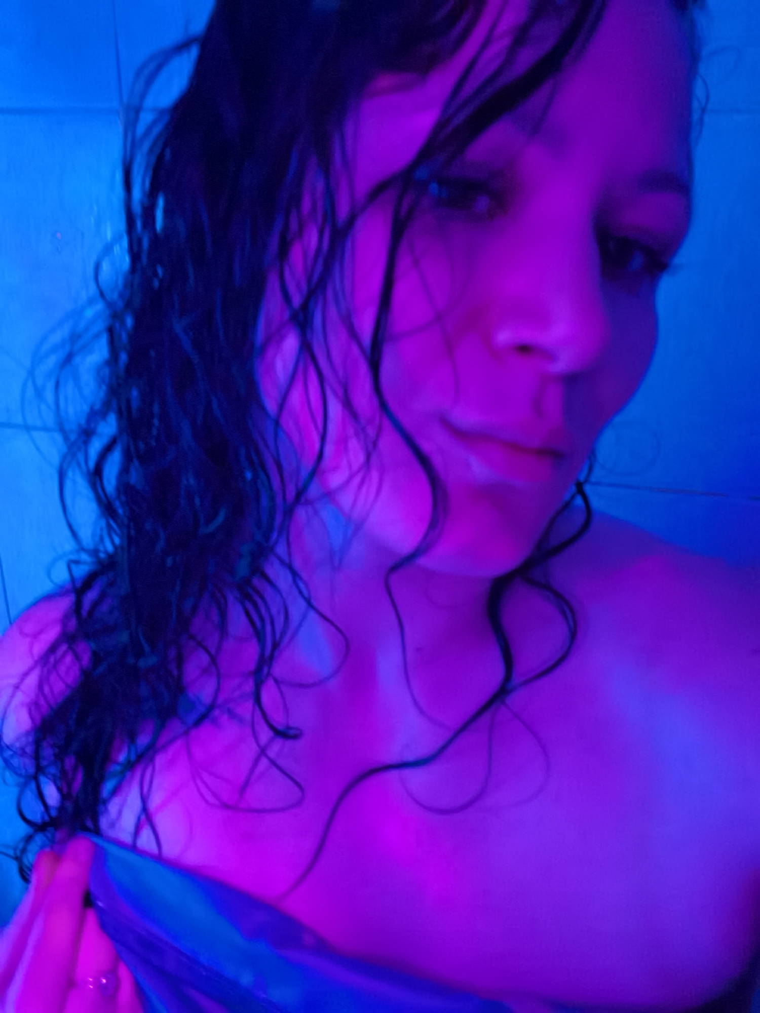A close-up shot depicts a person with wet, dark hair. The lighting is primarily blue and purple. The person appears to be partially undressed, possibly draped with a blue and purple fabric. The background consists of blue tiles. The city of Chaikovsky, Russia, is not discernible in the image.