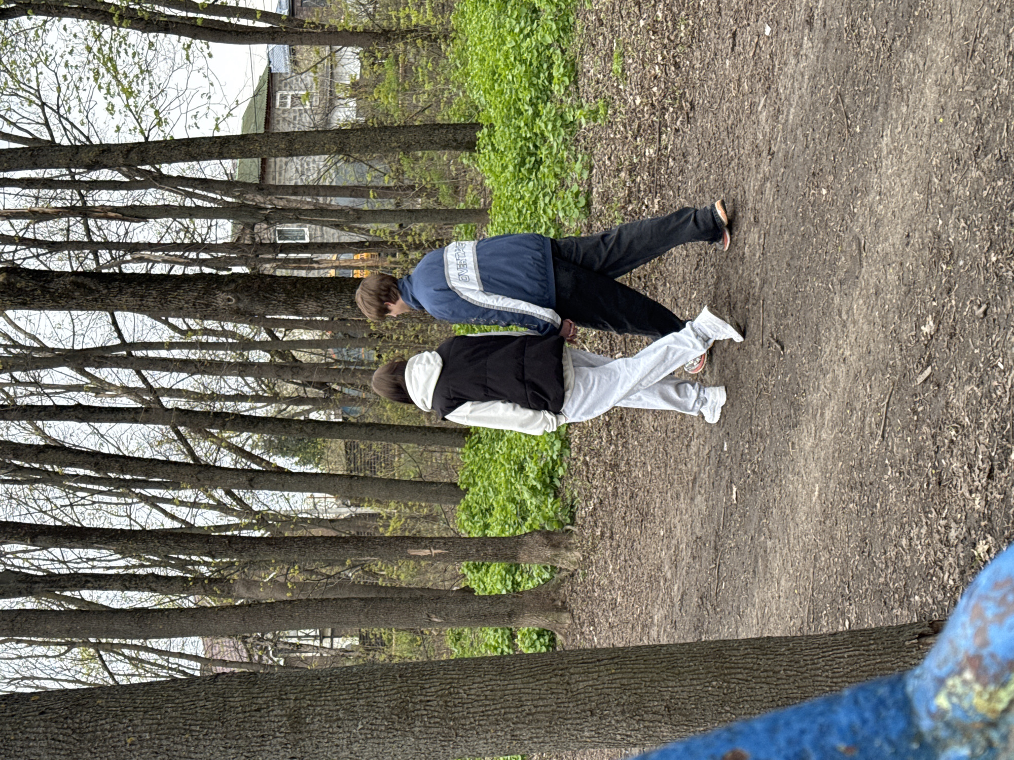 Two people walk along a dirt path lined with trees in a park setting. The person closest to the camera wears a black vest, a white hoodie, and light-colored pants with white sneakers. The other person is wearing a blue jacket with white accents and dark pants with darker footwear. The path is earthen, with scattered leaves and debris. Trees line the path on one side, with a small building visible in the background. The setting appears to be in a park or wooded area, likely located in Rostov-on-Don, Russia, based on the image request. There is a blue structure in the foreground.