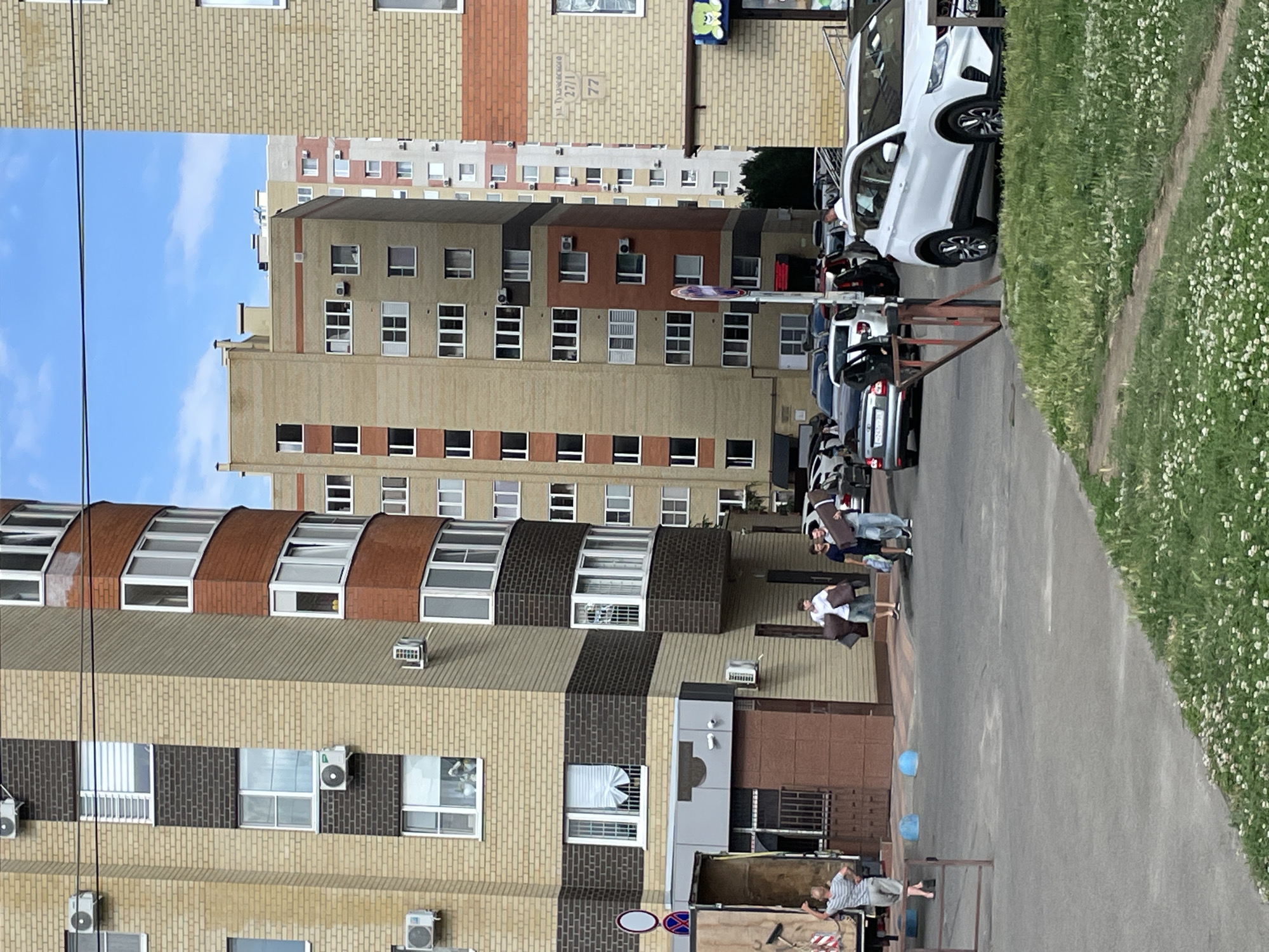 In the city of Stavropol, Russia, on a sunny day, three men are walking together on a paved area between apartment buildings. The man on the left wears a white t-shirt and shorts, carrying two large brown packages. The man in the middle wears a dark blue t-shirt and light-colored shorts, also carrying packages. The man on the right wears a grey t-shirt and dark pants, carrying similar packages.
To their left, a large truck is parked, with its rear doors open. A person in a striped t-shirt and shorts stands near the truck, facing away from the camera with their right arm raised, appearing to be directing or observing.
Several cars are parked and driving on the paved area. A white SUV is prominently parked on the right side of the image. In the background, multiple multi-story apartment buildings with brick and plaster facades are visible under a clear blue sky with some clouds. The buildings feature numerous windows, air conditioning units, and balconies. Some of the buildings have street numbers visible, including one with "ул. Тухачевского 27/1 77". A patch of green grass with small white flowers is present in the foreground.