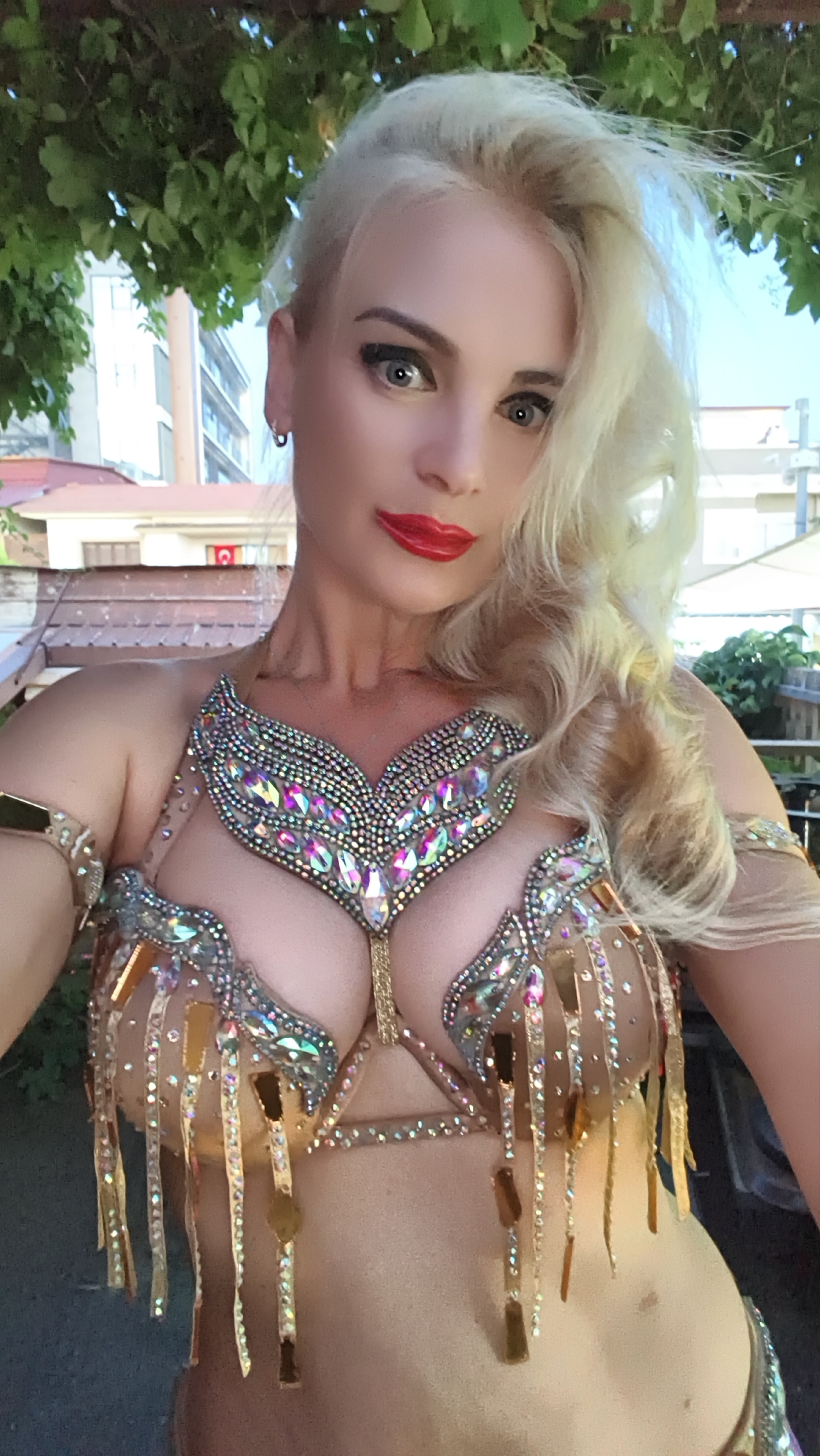 The image shows a woman with blonde hair, wearing a sparkling gold belly dancing costume with a bra adorned with rhinestones. She has bright red lipstick on and dark eye makeup. The woman is outdoors, with lush green foliage in the background and what appears to be buildings and rooftops behind her, suggesting an urban or semi-urban setting. There are also some trees and possibly a pergola structure visible. The lighting suggests it is daytime, with bright sunlight. The woman's expression is a direct gaze towards the camera, and her pose suggests she is ready for or in the midst of a performance. There are no visible text elements in the image. The location context is Kuşadası, Turkey.