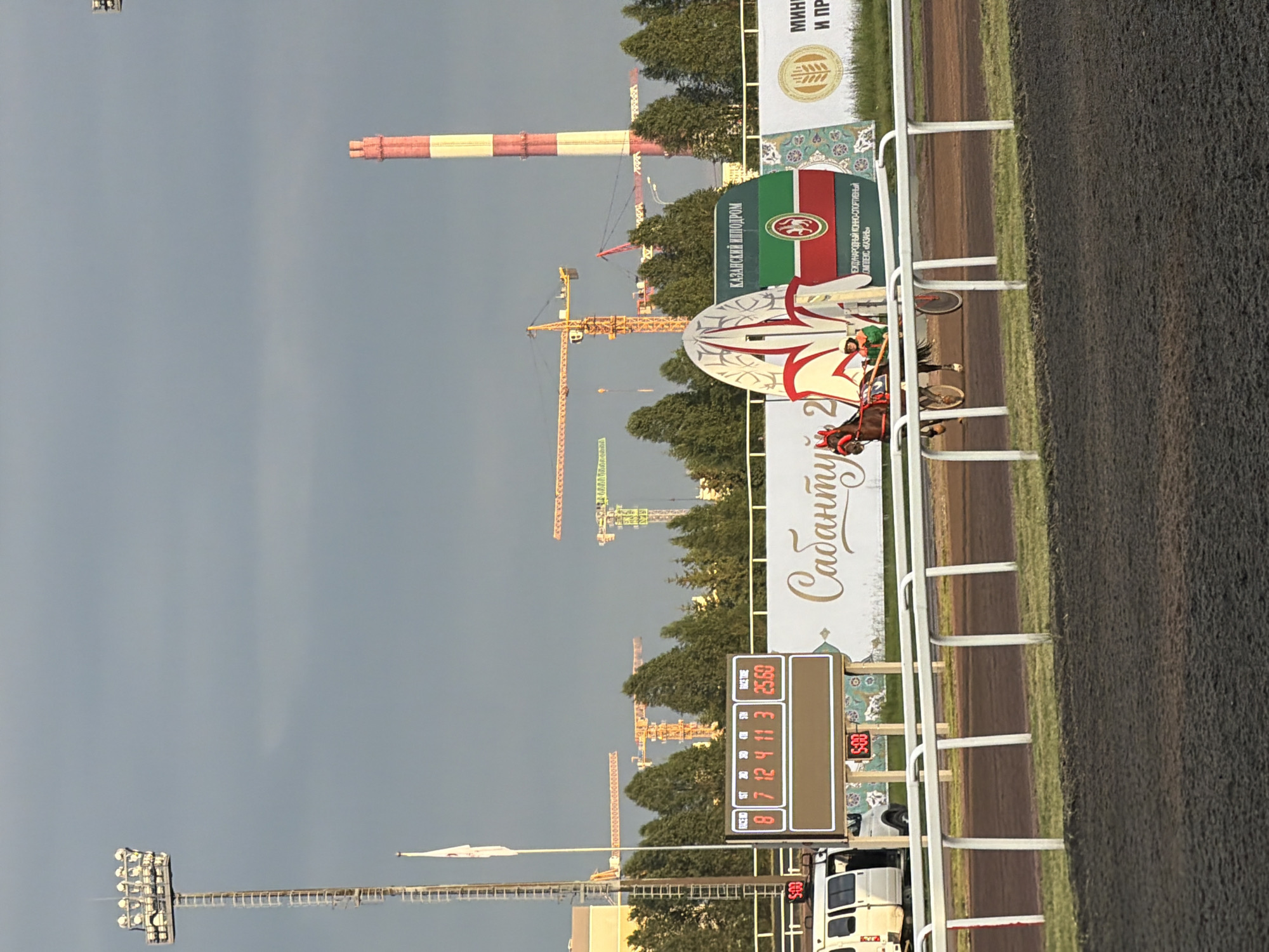 An outdoor, slightly overcast, daylight scene captures a horse-drawn cart participating in a race on a dirt track. A white railing delineates the track from the spectator area. In the background, a large, brightly lit digital scoreboard displays race information, including numbers "8 7 12 4 11 3" and a time of "25.60". Two smaller digital timers on poles read "5:00". A white van is parked near the scoreboard. Further in the background, a procession of construction cranes are visible against a hazy sky, indicating ongoing development. A tall, red and white striped smokestack also stands prominently. A large banner features text that appears to be in Russian and an image of a flag with a circular emblem. A horse with a red bridle and a driver in a green outfit are the primary subjects in motion on the track. The overall environment suggests a sporting event or a training session at a racetrack with significant industrial or construction activity nearby.