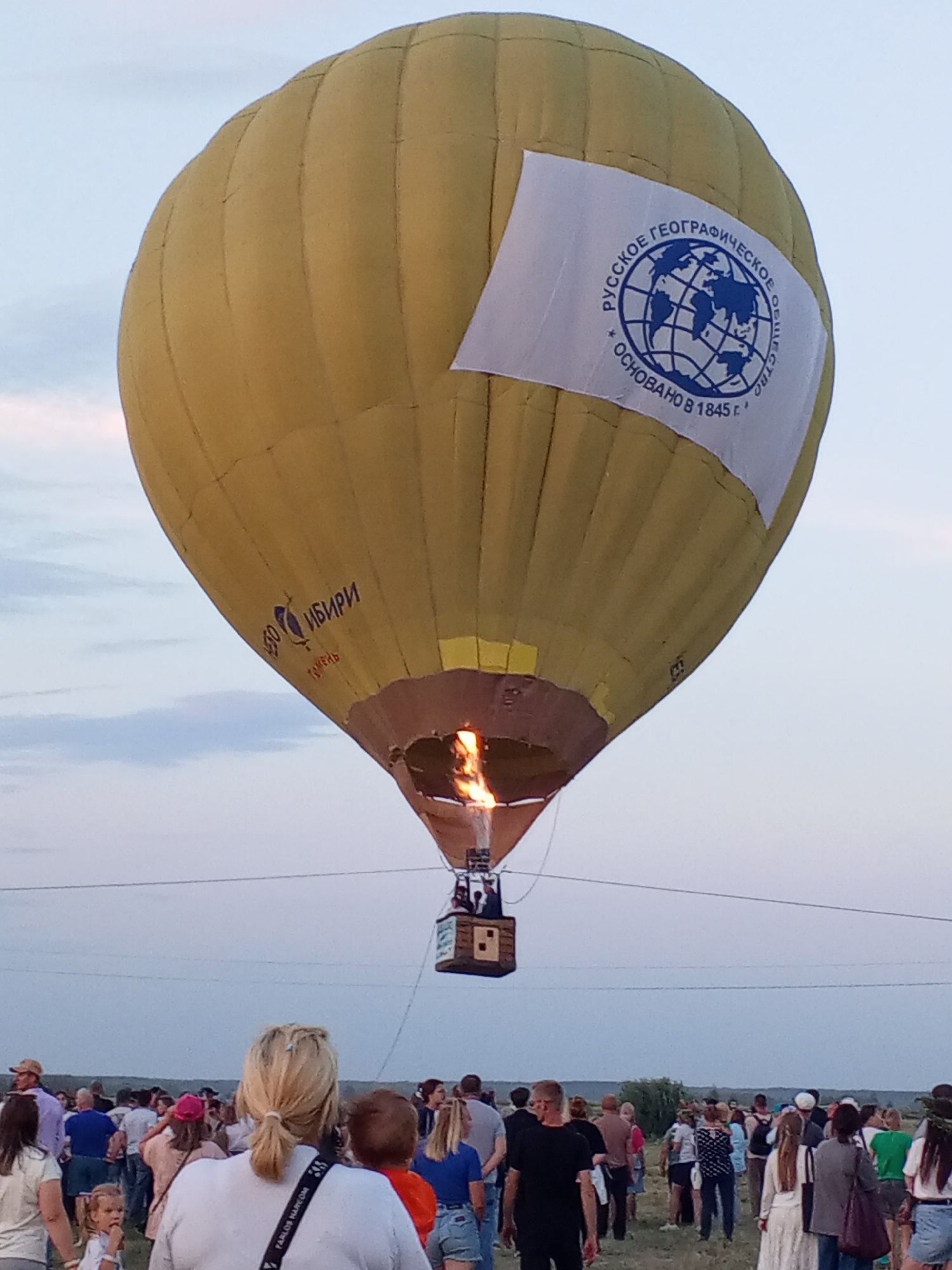 A bright yellow hot air balloon with a white banner featuring a blue globe and Russian text ascends into a pale sky. The banner reads "РУССКОЕ ГЕОГРАФИЧЕСКОЕ ОБЩЕСТВО ОСНОВАНО В 1845 г." Below the balloon, flames are visible emanating from the burner. A basket, carrying at least one person, hangs beneath the balloon. On the balloon's surface, there are Cyrillic inscriptions. In the foreground and middle ground, a large crowd of people stands on a grassy field, facing the balloon, suggesting an event or festival. The sky is overcast with light clouds. The horizon line is visible in the distance, showing a flat landscape with some greenery and a few indistinct structures.