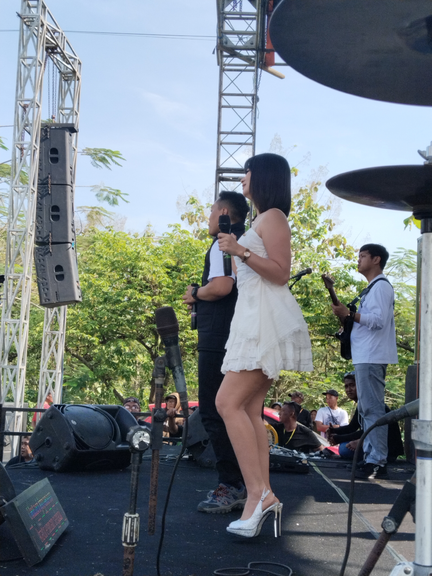 On an outdoor stage in Karanganom, Indonesia, a band performs for a crowd. A woman in a white dress and silver high heels stands near the center, holding a microphone and looking to the left. A man to her left holds a microphone and wears a black vest over a white shirt. A guitarist stands to the right, wearing a white long-sleeved shirt and gray pants. Behind them, green trees form a backdrop. The stage is black, and a sound system is visible, along with lighting trusses. The crowd sits or stands in front of the stage.
