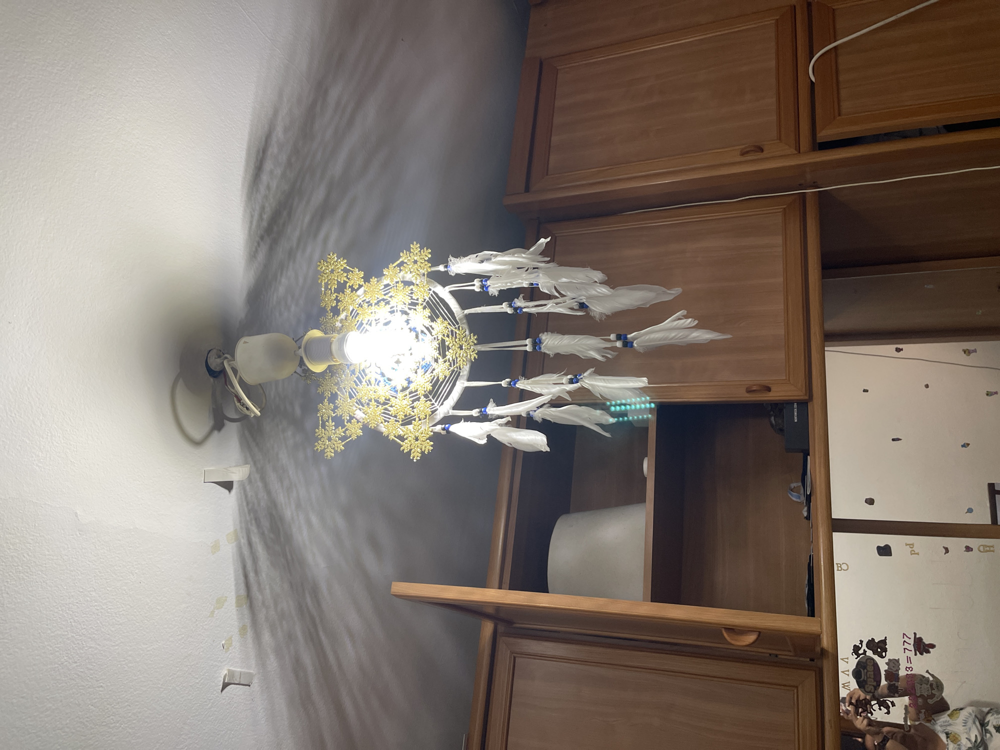 The image shows a ceiling light fixture with a dreamcatcher attached, hanging above a set of wooden cabinets. The light casts intricate shadows on the white ceiling. The cabinets appear to be part of a kitchen or storage area. The dreamcatcher has white feathers and blue beads. The scene is indoors, likely in a residential setting in Santa Coloma de Gramenet, Spain, though no specific landmarks or indicators of this location are visible. There are no people present in the image.