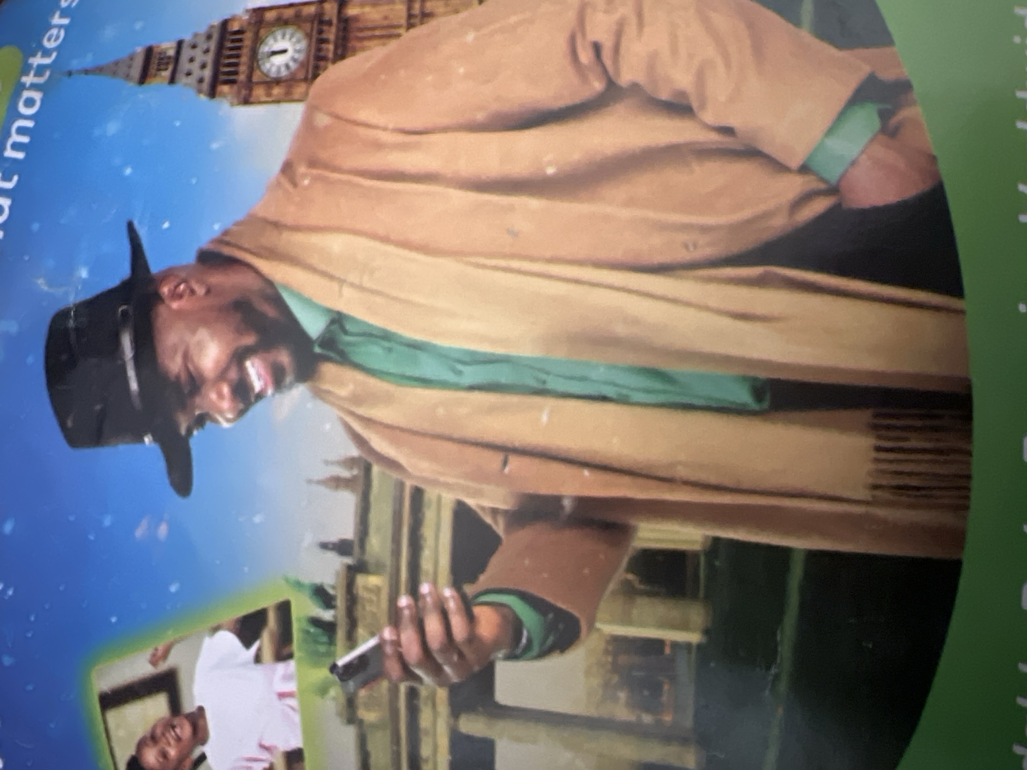 The image is a medium shot of a man wearing a black fedora, a green collared shirt, and a tan coat with a matching scarf. He is smiling and looking down at a smartphone he is holding in his left hand. In the background, to the left, there is a smaller inset image of a young girl wearing a white t-shirt. Behind the man, the iconic Big Ben clock tower is visible, suggesting a London setting. The overall impression is that this is an advertisement or promotional material, possibly for a service that connects people internationally, given the juxtaposition of the man in London with the inset of the child. There is no visual information present that would allow for identification of Jalingo, Nigeria.