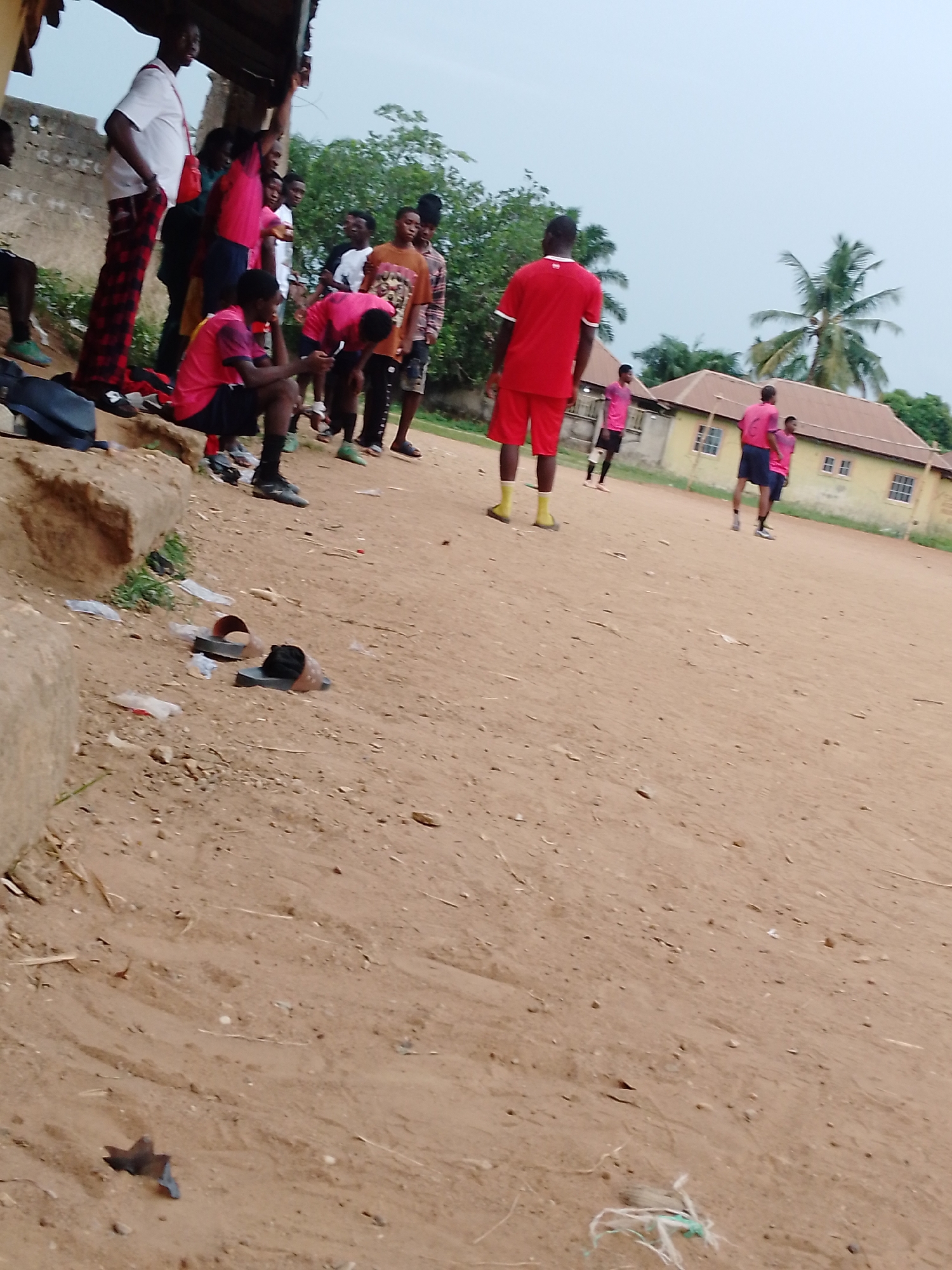 The scene is a dirt field in Unknown, Unknown, with a group of people gathered near a low building with a roof overhang to the left. Some of the people are wearing pink sports jerseys, while one is wearing a red jersey. Several people are on the field, with a building behind them with shuttered windows. Two sandals are discarded on the ground near the edge of the field. A palm tree is visible in the background.