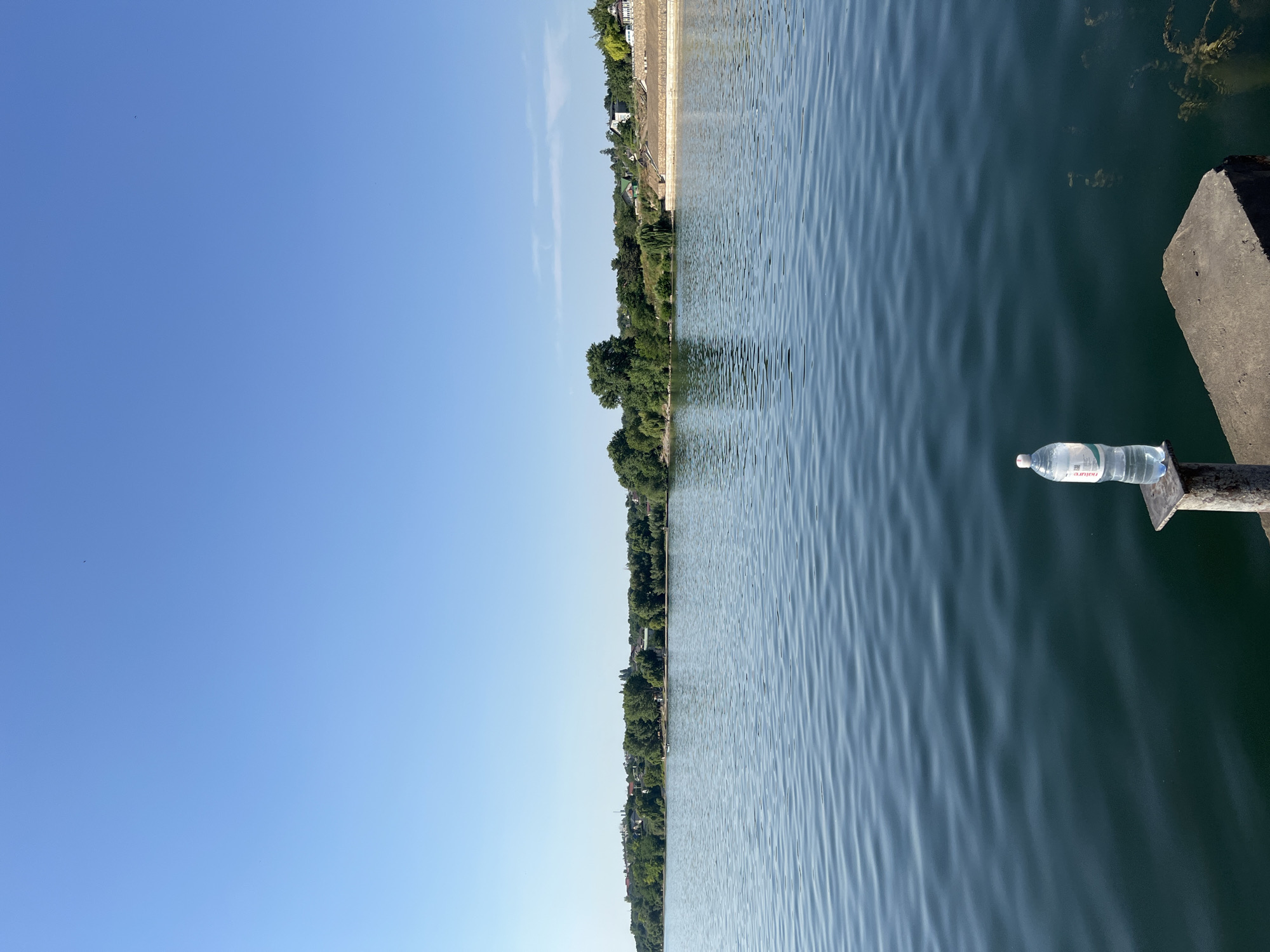 This is a vertical, medium-shot, eye-level photograph taken from a pier or dock overlooking a body of water. In the foreground, on a metal post and a concrete slab, sits a clear plastic bottle of water with a white cap. The water in the lake is dark blue and rippled by a gentle breeze. The far bank of the lake is lined with a dense row of green trees, behind which a few buildings and rooftops are visible. The sky is clear, bright blue, and cloudless, indicating a sunny day. The overall scene depicts a serene natural environment with a touch of man-made elements, likely in the city of Korzhevo, Moldova, given the context. There are no people visible in the frame.