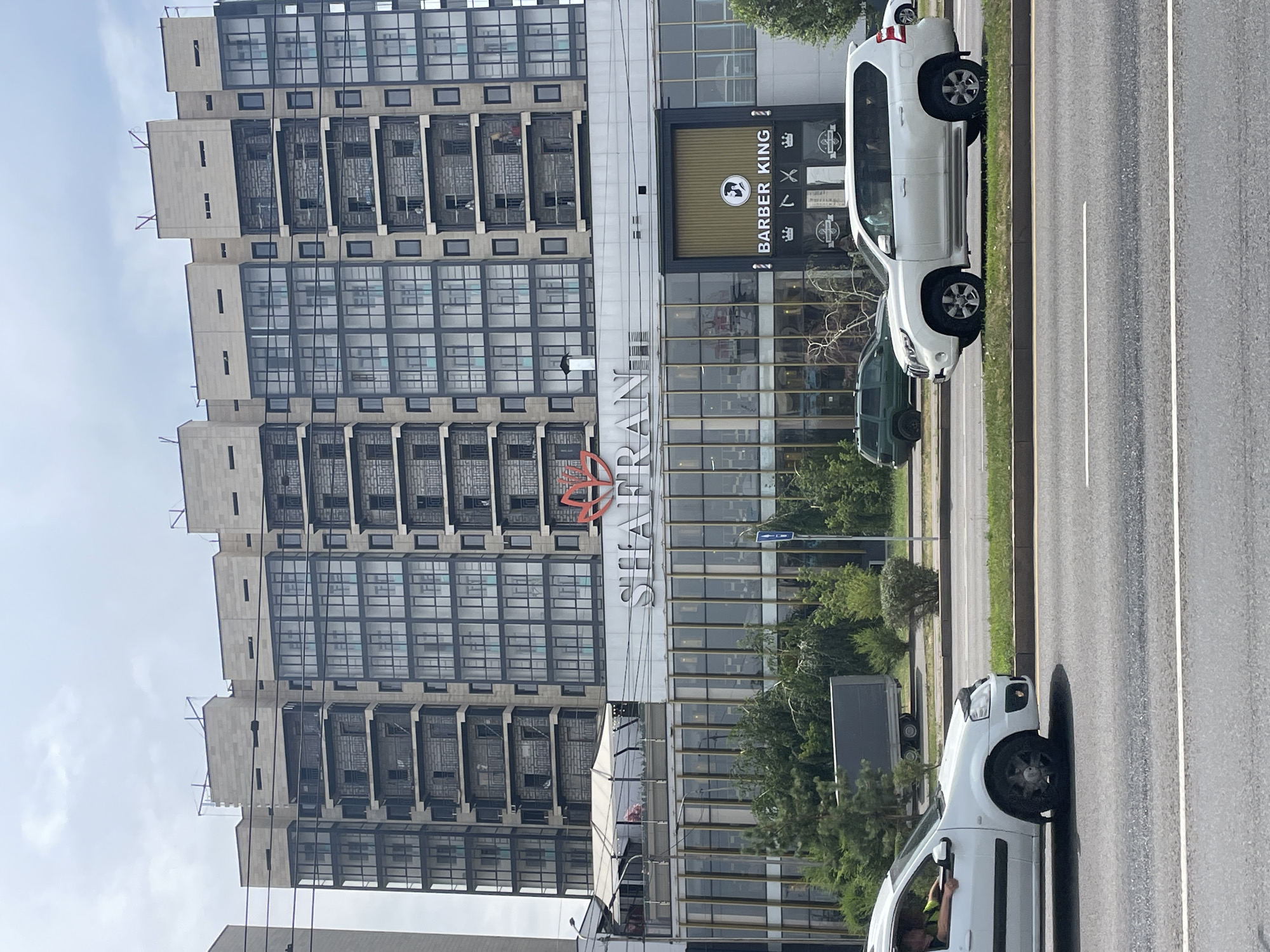 The image shows a modern, multi-story building with a commercial ground floor. The building is clad in light-colored stone and features numerous glass-enclosed balconies on its upper levels. The ground floor has a large signage displaying "SHAFRAN" in prominent white lettering, accompanied by a red stylized floral logo. Adjacent to this, there is a storefront with the words "BARBER KING" displayed above its entrance, which is framed by gold-colored accents. The street level in front of the building shows a road with passing vehicles, including a white sedan and a white SUV. Several trees and green spaces are visible between the road and the building. The sky is overcast, suggesting a cloudy day. The architectural style and signage suggest an urban commercial and residential area. The presence of "Shara" and potentially local businesses indicates this location is within Almaty, Kazakhstan.