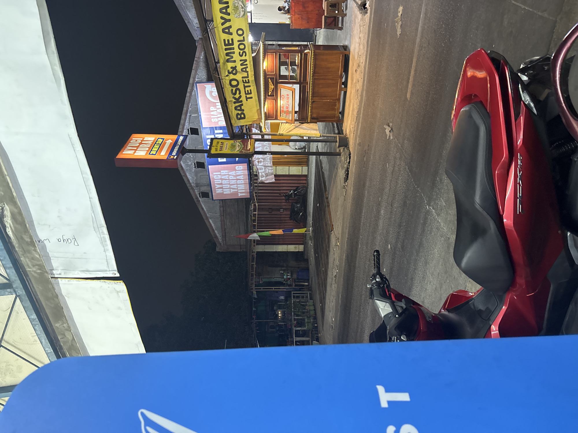 The image depicts an outdoor scene at night, likely a street-side food stall area in North Jakarta, Indonesia. A red scooter is parked in the foreground, partially obscured by a large blue structure. In the background, several food stalls are visible. One stall features a yellow banner with Indonesian text that translates to "BAKSO & MIE AYAM TETELAN SOLO," indicating it sells meatballs and chicken noodles from Solo. Another stall has a sign advertising "NYUCI MURAH" (cheap laundry) and "GEMBIRA LAUNDRY." The environment appears to be a bustling commercial area with some trees and buildings visible in the background. The lighting suggests it is nighttime, with artificial lights illuminating the stalls and the street.