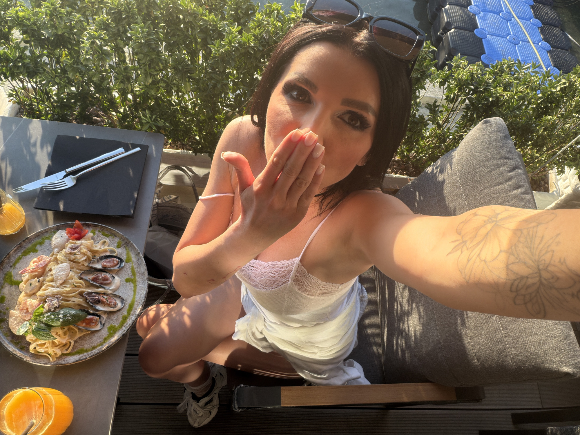 A woman takes a selfie at an outdoor restaurant in Rafailovići, Montenegro. She is sitting at a table with a plate of seafood pasta, cutlery, and orange juice. She wears a white dress and has sunglasses on her head. The background shows green foliage and a floating platform on the water.
