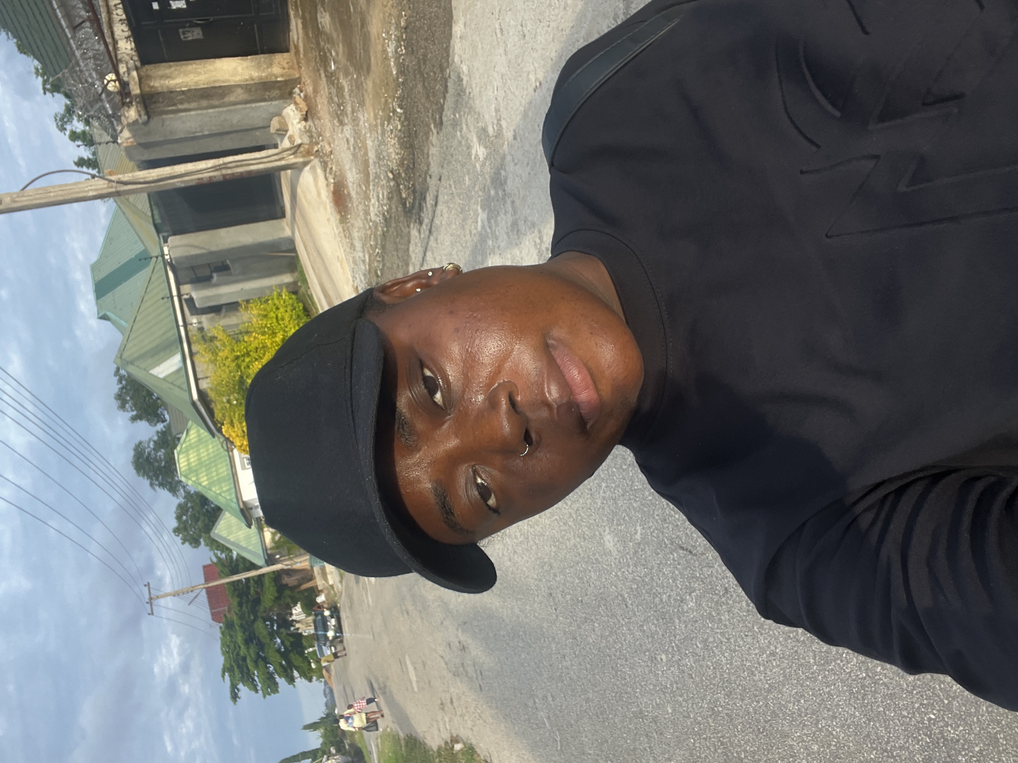 This image captures a street scene in Abuja, Nigeria, featuring a prominent selfie in the foreground.
In the foreground, a person with dark skin is taking a selfie, looking directly at the camera with a neutral expression. They wear a black baseball cap and a long-sleeved black shirt with a dark, embossed design on the chest, possibly a stylized letter. A silver hoop nose ring adorns the left nostril, and two small earrings (one stud, one hoop) are visible on the left earlobe. A black strap, likely from a bag, crosses their right shoulder. The lighting highlights the right side of their face and shoulder, causing a slight sheen on the skin.
The middle ground reveals a grey asphalt road, appearing somewhat worn. A concrete utility pole stands on the right side of the road, supporting multiple overhead power lines that stretch across the frame. Green trees and shrubs line the road, with a bright yellow flowering bush visible on the right.
In the background, the road curves gently to the left. Several residential buildings with concrete walls and prominent green-colored pitched roofs are visible on both sides of the street. Some properties feature high walls, with razor wire or barbed wire visible atop a wall on the right. Further down the road on the left, two individuals, an adult in a light-colored outfit and a child, are walking away from the camera. A dark-colored vehicle is also visible in the distance on the left side of the road. The sky overhead is largely overcast with grey clouds, indicating daytime conditions.