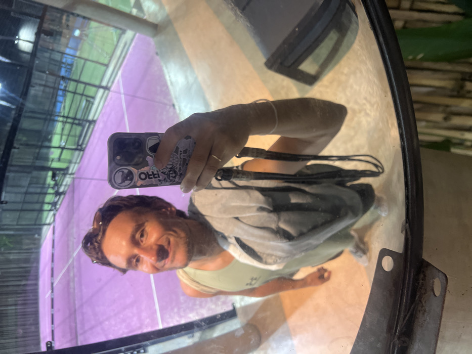 A male individual is depicted in a mirror selfie within a padel tennis facility located in Pecatu, Indonesia. The subject, a Caucasian male with brown hair, is smiling and wears sunglasses positioned on top of his head. He is dressed in a light green tank top, bearing a faint logo, and light-colored shorts and sneakers are partially visible. A gray garment is draped over his left shoulder. He holds a dark-colored smartphone, adorned with multiple stickers including a bull's head logo and text "OFF!", in his right hand to capture the image. A thin, metallic ring is visible on his right ring finger, and a light-colored bracelet is on his right wrist. The background, reflected in the curved mirror, displays a brightly lit padel court with a distinct purple playing surface and white boundary lines. The court is enclosed by transparent glass walls and metal mesh fencing. A second padel court, featuring a green playing surface, is visible in the deeper background, suggesting a multi-court complex. The mirror itself has a dark metal frame along its lower edge, and a vertical bamboo-like structure is present in the unreflected portion of the scene to the bottom right.