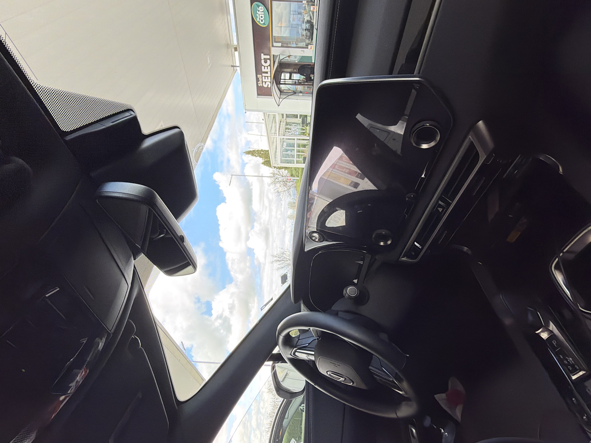 The image, taken from inside a car, shows the view through the windshield toward a Shell Select and Shell Cafe gas station. The sky is partly cloudy. The car's interior is primarily black, with the steering wheel and dashboard prominently visible. The location appears to be in the city of Dragor, Bulgaria, based on the signage of the Shell gas station. There are no people visible inside the car. A person can be seen entering the cafe.