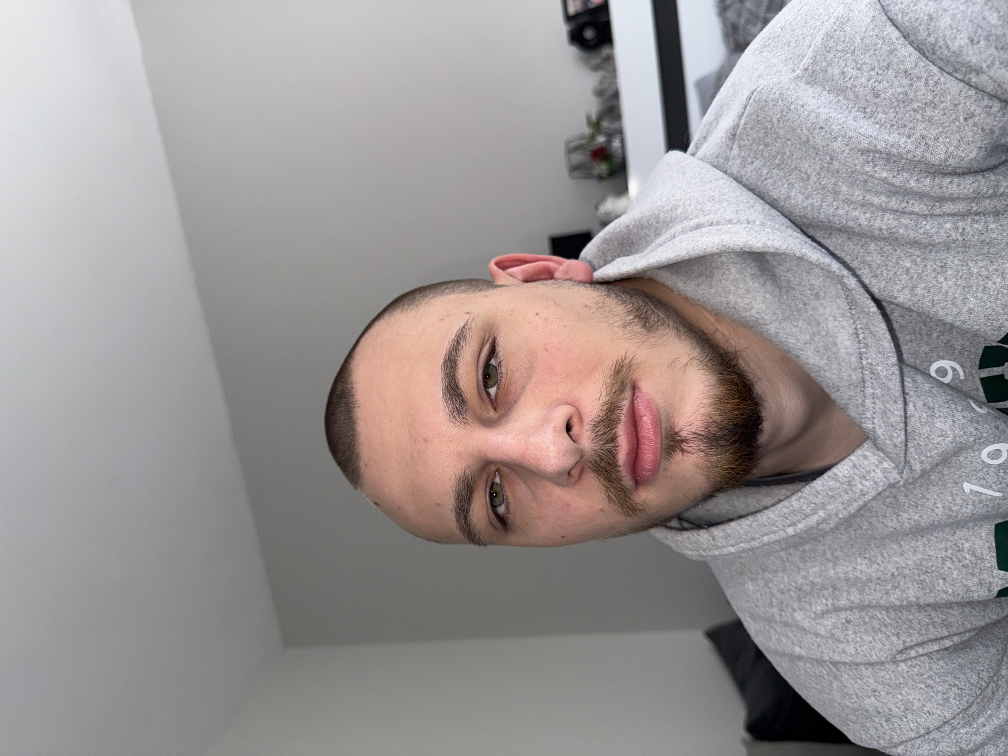 A man with a shaved head and short brown beard is wearing a grey hooded sweatshirt. The photo appears to be taken from a low angle looking up at the man. The background includes a white wall and a glimpse of a dark-colored object and decorative items on a shelf. The image does not contain any location information related to Stara Zagora, Bulgaria.