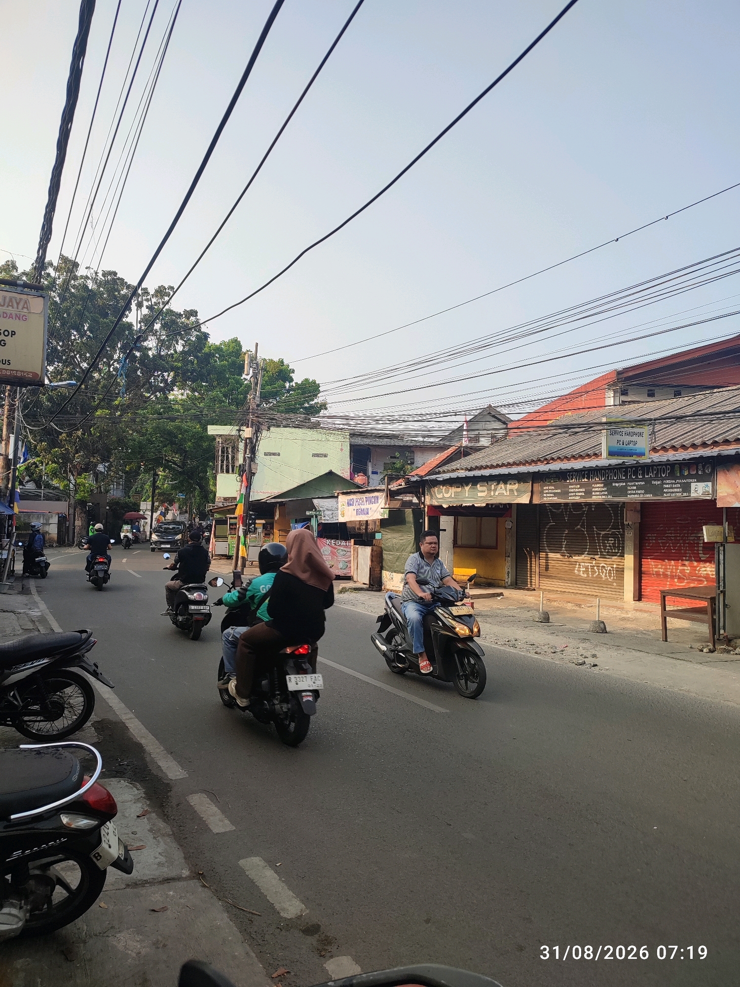 The image depicts a street scene in East Jakarta, Indonesia, captured at 07:19 on August 31, 2026. The street is asphalt, with faded white lane markings. Several motorcycles are present on the road. One motorcycle in the foreground, on the left side, is partially visible. Another black motorcycle is parked on the sidewalk on the far left. Further down the street, a group of three people ride a motorcycle; the rider in the front is wearing a green shirt and helmet, and the two passengers behind are wearing a blue shirt and jeans, and a brown hijab and long-sleeved shirt respectively. Another motorcycle with a single rider, wearing a blue shirt, jeans, and a helmet, is moving in the same direction. A third motorcycle is further up the road with a rider wearing a grey t-shirt and shorts. A black car is also visible in the distance.
On the right side of the street, a row of buildings with tiled roofs line the road. One building displays signage that reads "COPY STAR" and "SERVICE HANDPHONE PC & LAPTOP". The shutters of these establishments are down, with graffiti visible on some. A wooden table and chair are placed outside one of the buildings. On the left side of the street, more buildings and trees are visible. The sky is clear with a pale blue hue. Overhead, a complex network of electrical wires is strung across the street. The lighting suggests it is early morning.