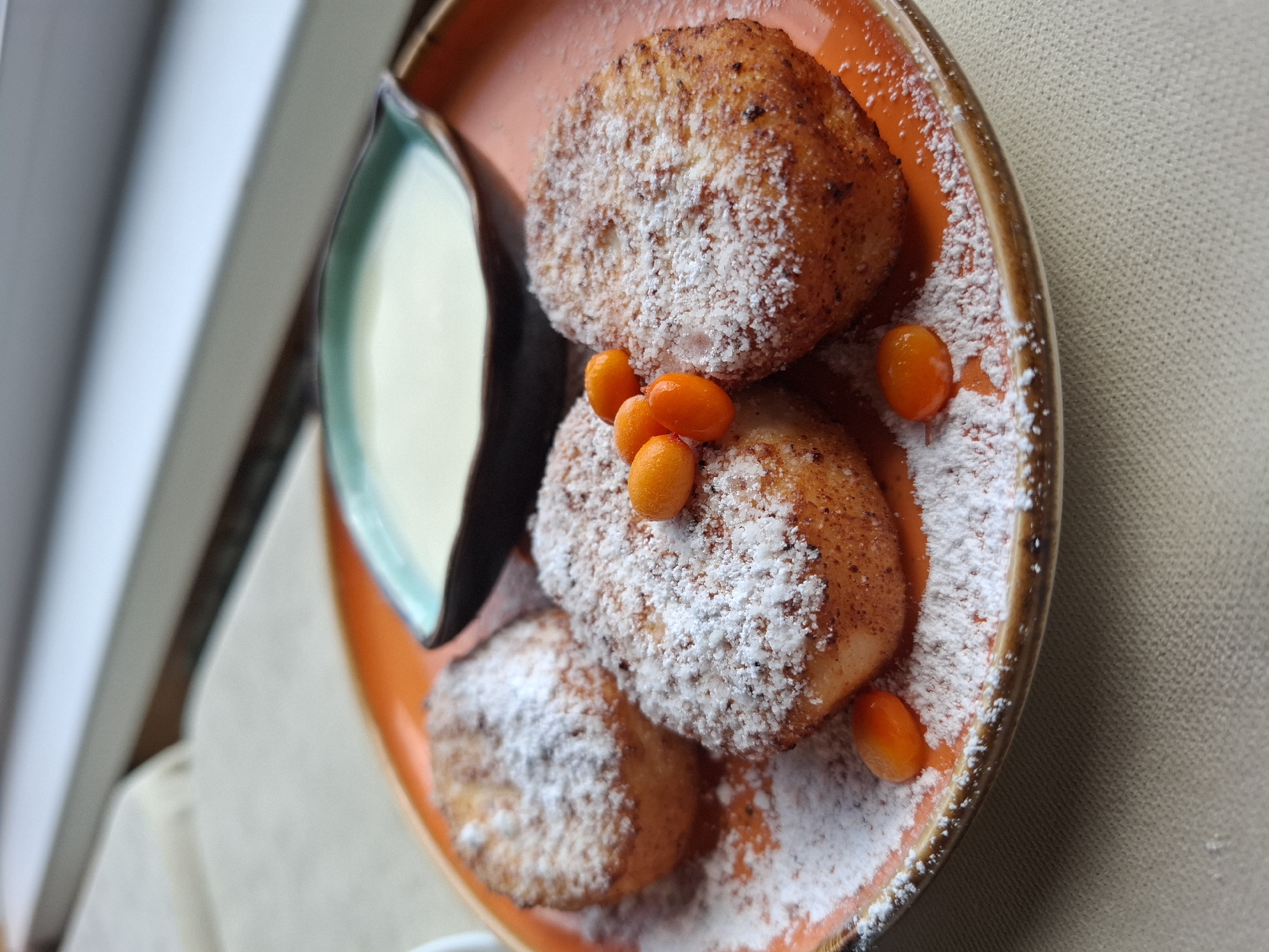 The image shows a plate of syrniki, a type of fried curd cheese pancake, dusted with powdered sugar and garnished with sea buckthorn berries. A small dish of white sauce, likely sour cream or a sweetened condensed milk, is placed behind the syrniki. The plate is orange and sits on a beige tablecloth. The background is blurred, showing what appears to be a windowsill with a view of a building. The scene is lit from the side, casting soft shadows. There are no people or actions depicted in the image, and the location in Sukhoy Ruchey, Russia, is inferred from the context and not visually evident.