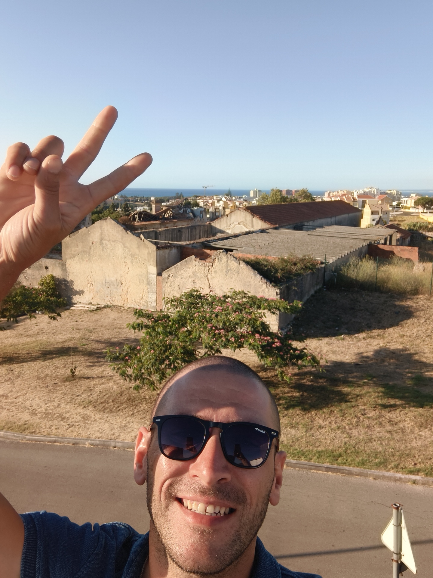 A man with a shaved head and sunglasses smiles at the camera, holding up two fingers in a peace sign. He is wearing a blue polo shirt. In the background, there are dilapidated buildings, dry grass, and a view of the city of Cascais, Portugal, with the ocean in the distance. The sky is clear and blue. The man is likely taking a selfie on a sunny day.