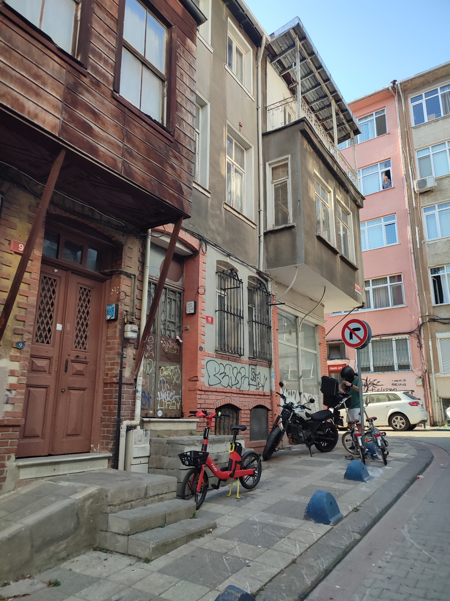 The image captures a narrow street scene in Kadıköy, Turkey, characterized by multi-story buildings with varying architectural styles and materials. On the left, a weathered, wooden-clad upper story with a projecting balcony overhangs a brick facade with a large, reddish-brown double door. Adjacent to it, a grey building features a narrow balcony with a metal-sheeted roof. Further right, a pink building stands next to a pale grey one, both with multiple windows. A woman is visible looking out from a window in the pink building.
On the street level, a red electric bicycle with yellow accents is parked near the entrance on the left. To the right, a man in a dark helmet and a green shirt is standing next to a black motorcycle with a black storage box on the back. A smaller bicycle is parked next to the motorcycle. A white car is visible parked further down the street on the right. A prominent "no right turn" traffic sign is positioned on a pole. The sidewalk is paved with grey concrete blocks, and blue, wedge-shaped bollards are placed along the curb. Graffiti is present on the walls of the buildings, particularly on the lower sections of the grey building and the brick area to the right of the double doors. The scene is illuminated by sunlight, suggesting it is daytime.
