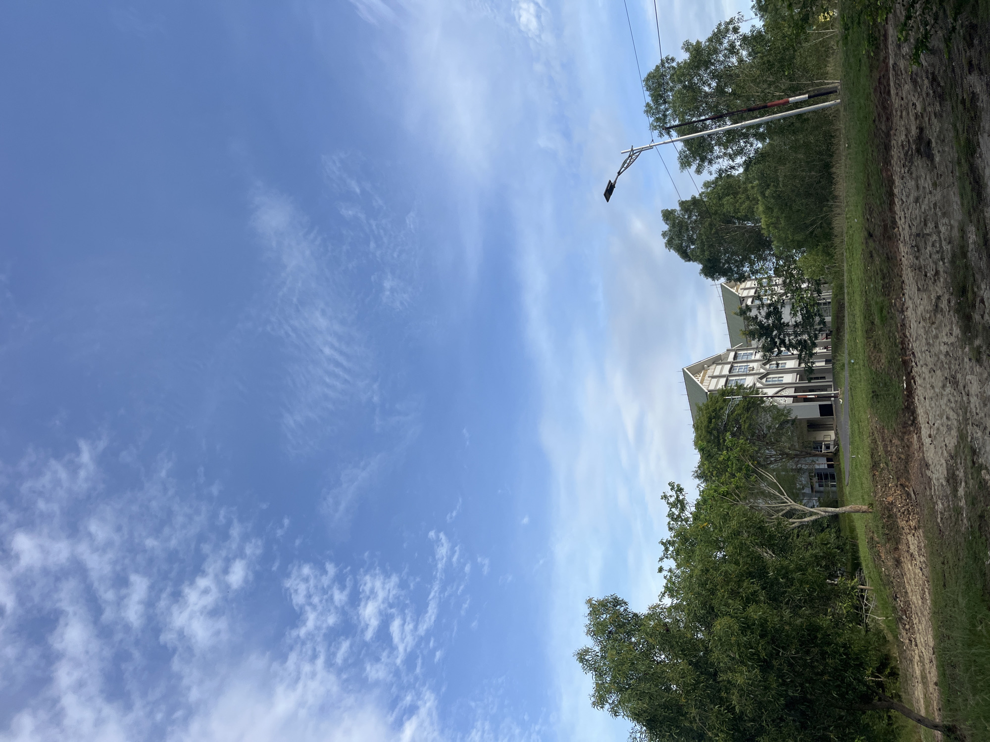 The image shows a sky with light clouds over Lhokseumawe, Indonesia. A white apartment building is visible behind trees and a light post. A dirt and grass path runs along the right side of the frame.