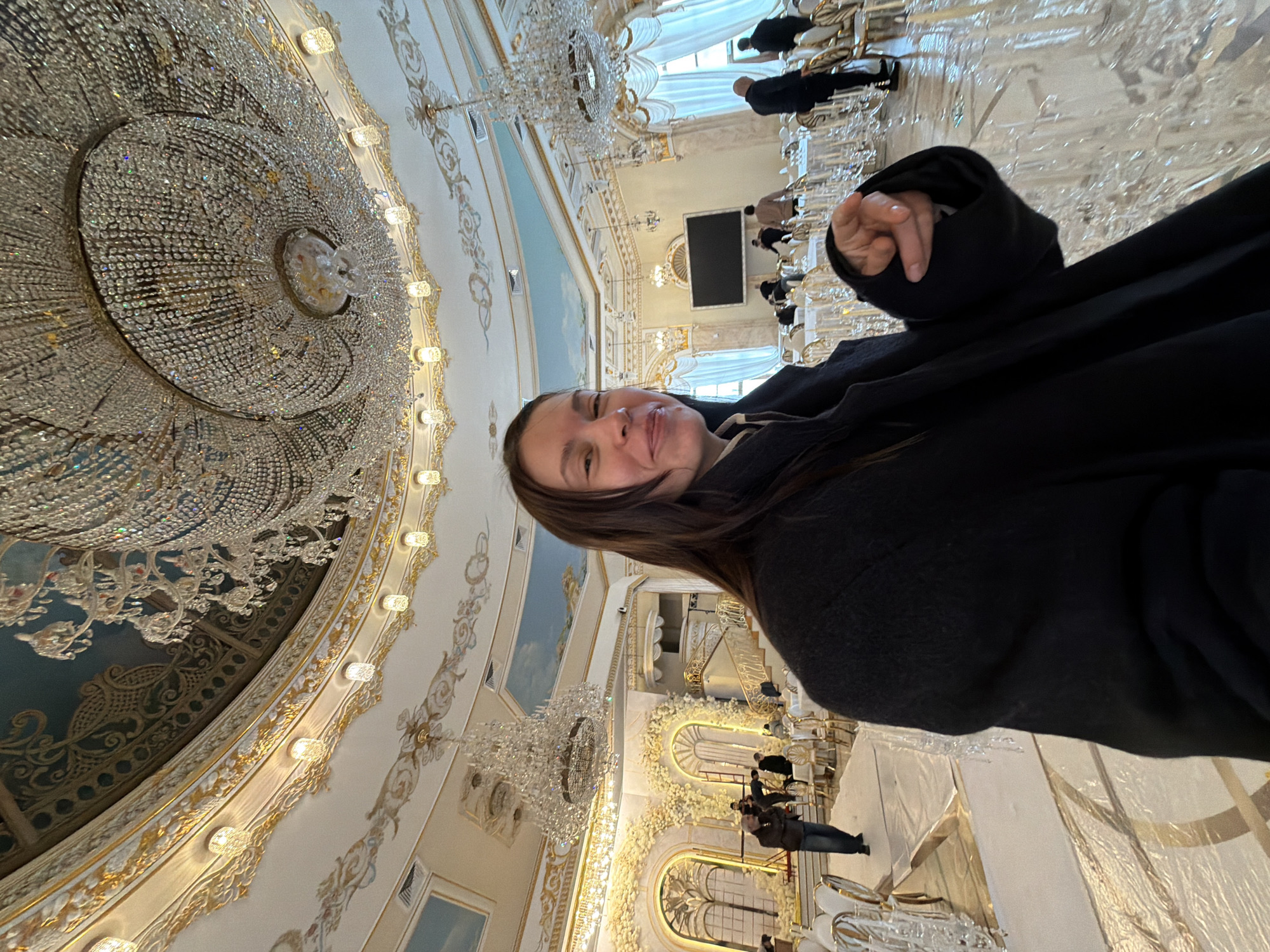 A woman wearing a dark coat poses for a selfie in a grand, ornately decorated hall. Above her is a massive crystal chandelier, and the ceiling is adorned with intricate gold detailing and painted scenes. In the background, other people can be seen walking around, observing the hall's architecture. Rows of white tables are set up with clear glassware. The overall impression is one of opulence and grandeur, and the location is likely the Winter Garden inside the State Historical Museum in Moscow, Russia.