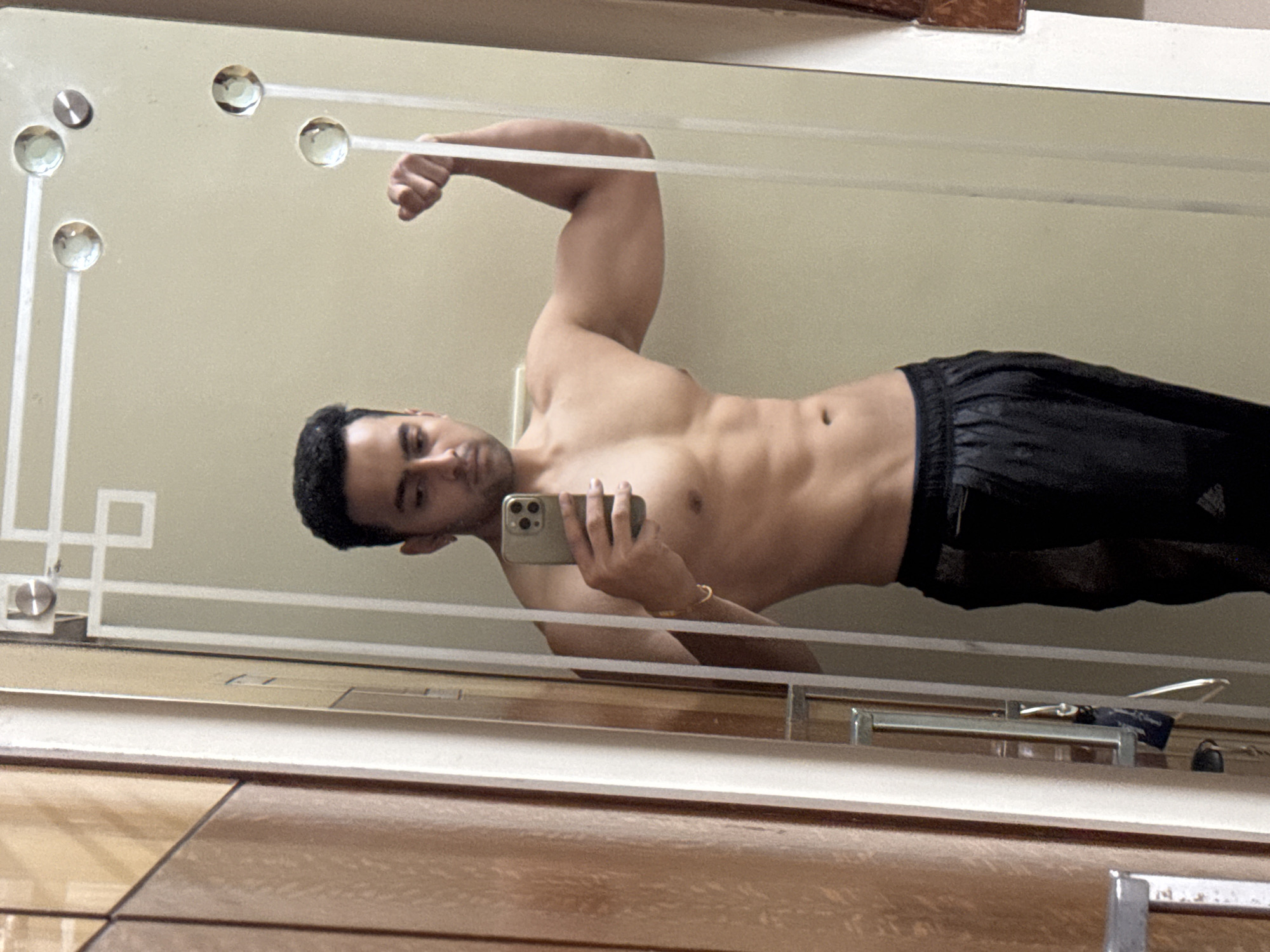 A man is taking a selfie in a mirror. He is shirtless, wearing black shorts. The background shows a light-colored wall and a wooden surface. The location is unknown, but cannot be confirmed as Kolkata, India.
