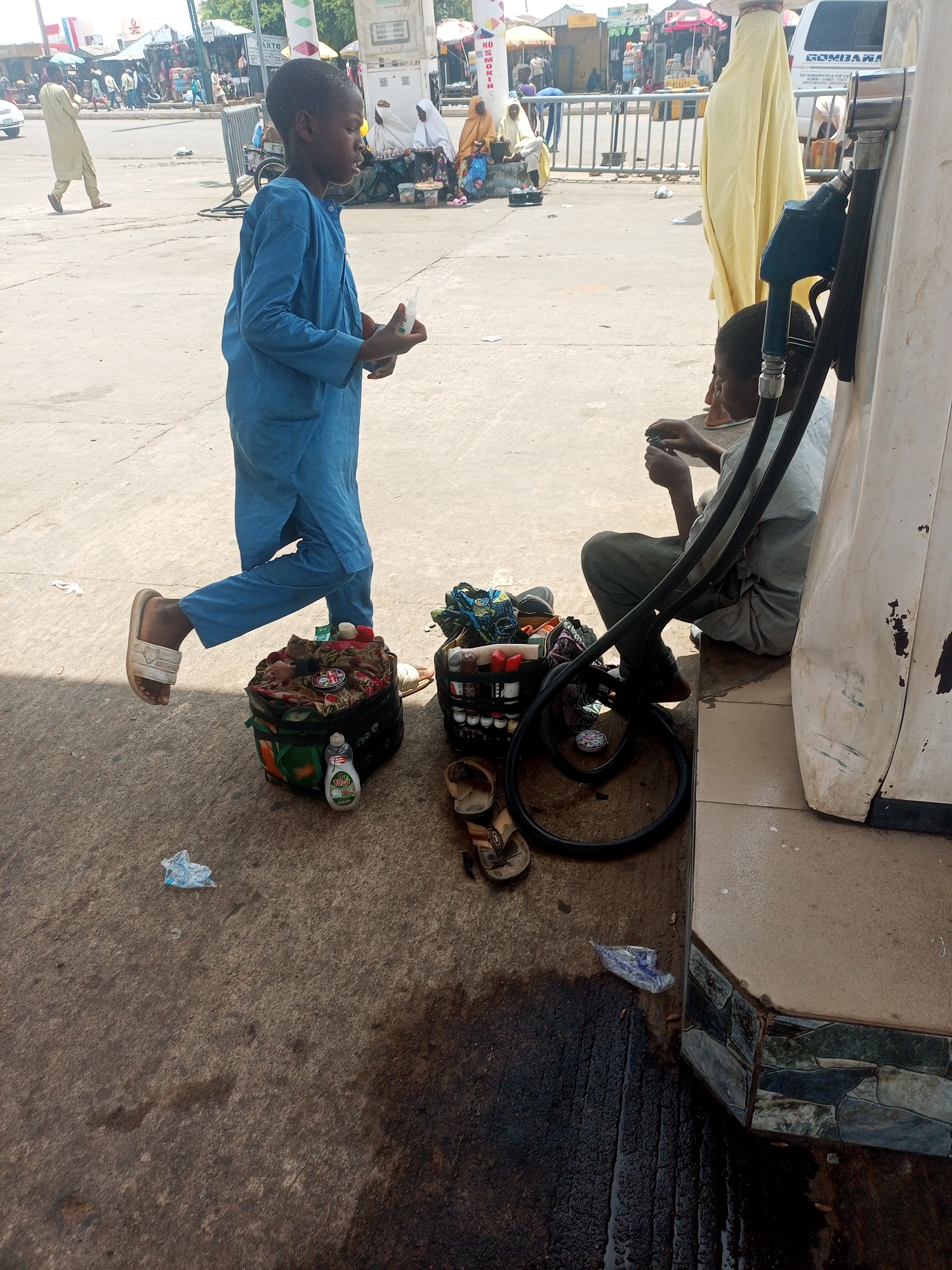 The image captures a scene in Gombe, Nigeria, featuring two young boys at what appears to be a roadside service station. One boy, dressed in a blue outfit and sandals, is standing and holding a small bottle. He is positioned next to a collection of bags and containers, likely containing his tools and materials. The second boy sits near a fuel pump. A tire rests near him. The surrounding area seems to be a busy street, with people and vehicles in the background.