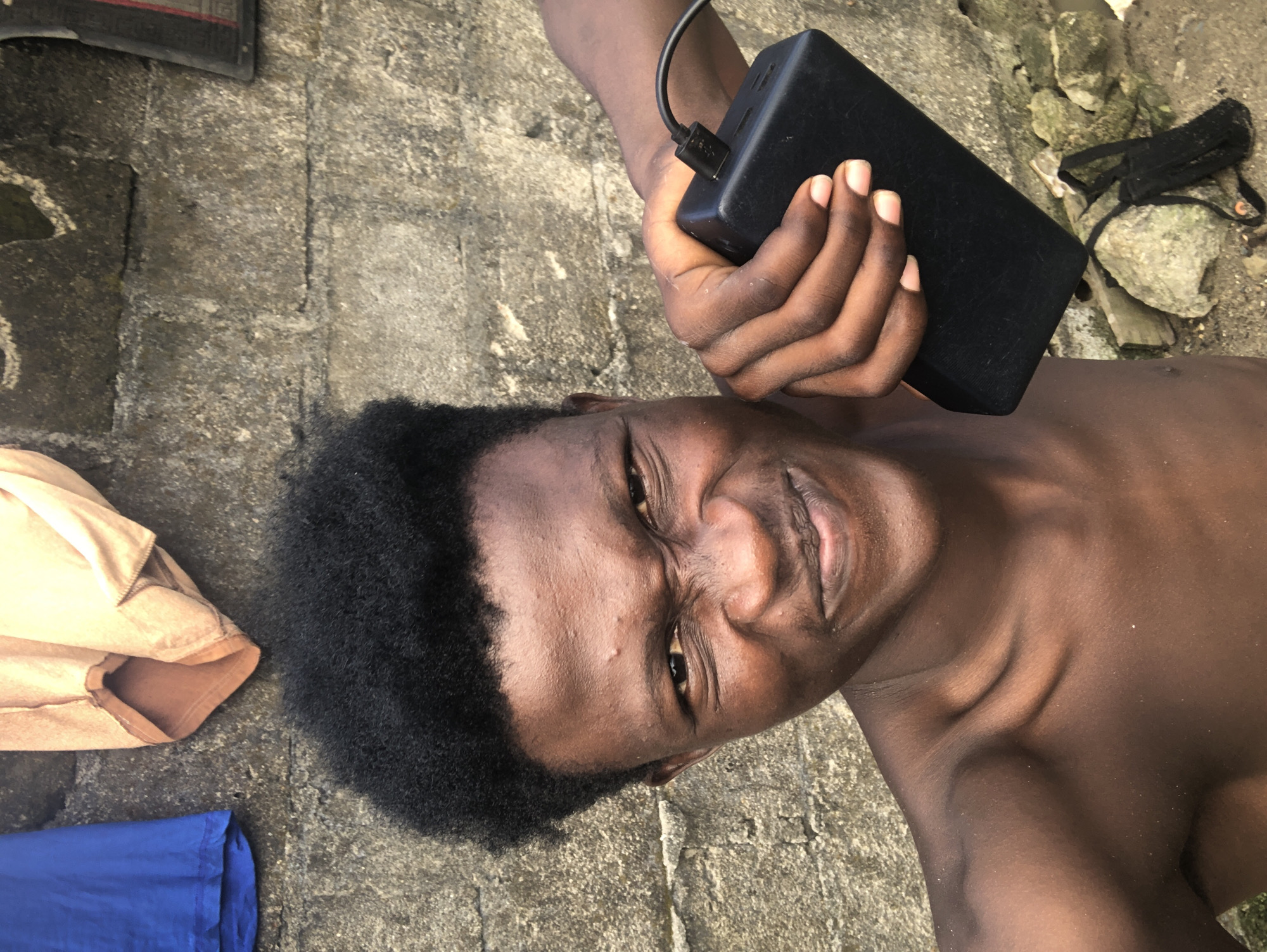 A close-up, chest-up shot of a young Black male in Ado, Nigeria. He is shirtless and facing the camera, with his left arm extended, holding a black portable charger with a cable plugged into it. The man has a strong, grimacing expression, with his eyebrows furrowed and his mouth slightly open, revealing his teeth. He has a dark, short afro. The background consists of a rough, grey concrete wall, and to the left, two pieces of fabric are hanging, one blue and the other light brown. The ground beneath the wall is covered in dirt and rocks, with a black fabric item lying on it. The lighting suggests it is daytime, with harsh shadows.