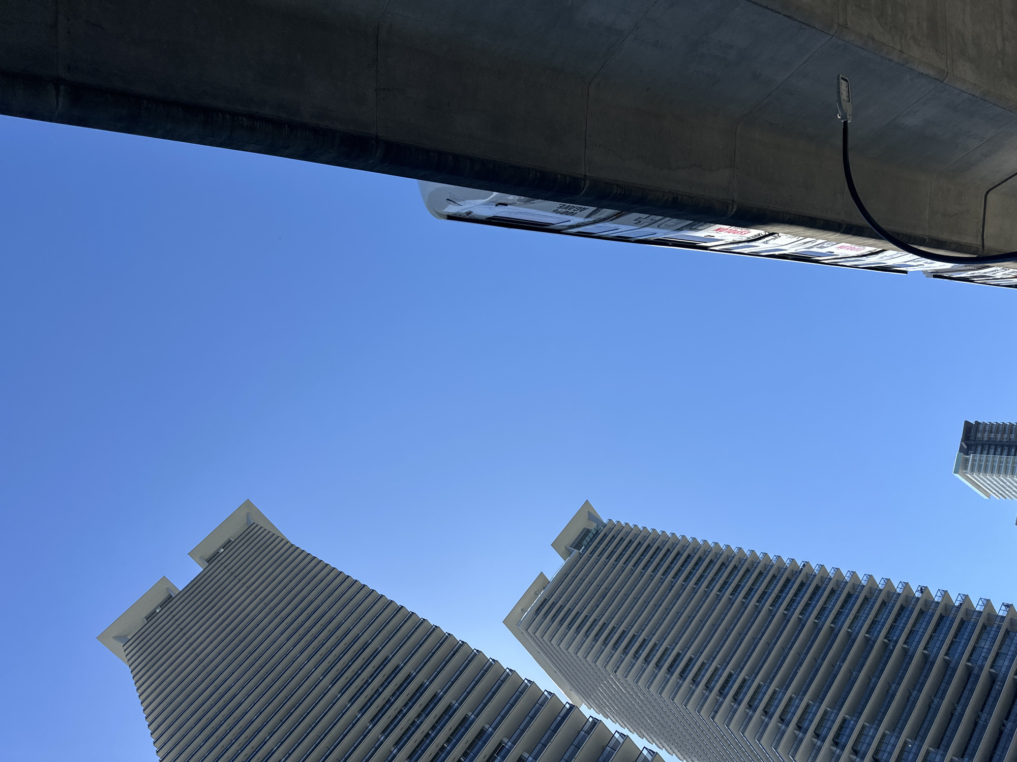 This is a low-angle, full-frame, vertical shot of two modern high-rise buildings against a clear, bright blue sky. The buildings are tall and slender with a repeating pattern of balconies and windows, giving them a textured, linear appearance. The building on the left is closer and larger in the frame, angled away from the viewer. A second, similar building is positioned behind and to the right of the first. In the foreground, on the right side of the image, a portion of a concrete overpass or elevated structure juts into the frame from the right. A street lamp with a curved arm extends from beneath this structure. A train, with visible advertisements on its side, is partially visible moving along the elevated track. The scene appears to be in an urban environment, likely a city, with Burnaby, Canada being the identified location. The sunlight is strong, casting distinct shadows on the understructure. There are no people visible in the image.