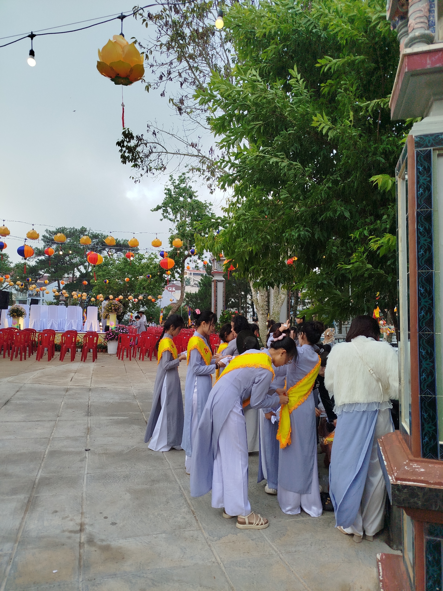 The image shows a group of young women dressed in traditional grey and white Vietnamese Ao Dai attire with yellow sashes, standing on a stone-tiled plaza. They are near the edge of a building with tiled decorations. In the background, there is an outdoor event setup with rows of red plastic chairs facing a white-covered stage or platform. Decorative hanging lotus lanterns and string lights are strung overhead. Trees and greenery are scattered around the plaza. The sky is overcast. This scene is located in Da Lat, Vietnam.