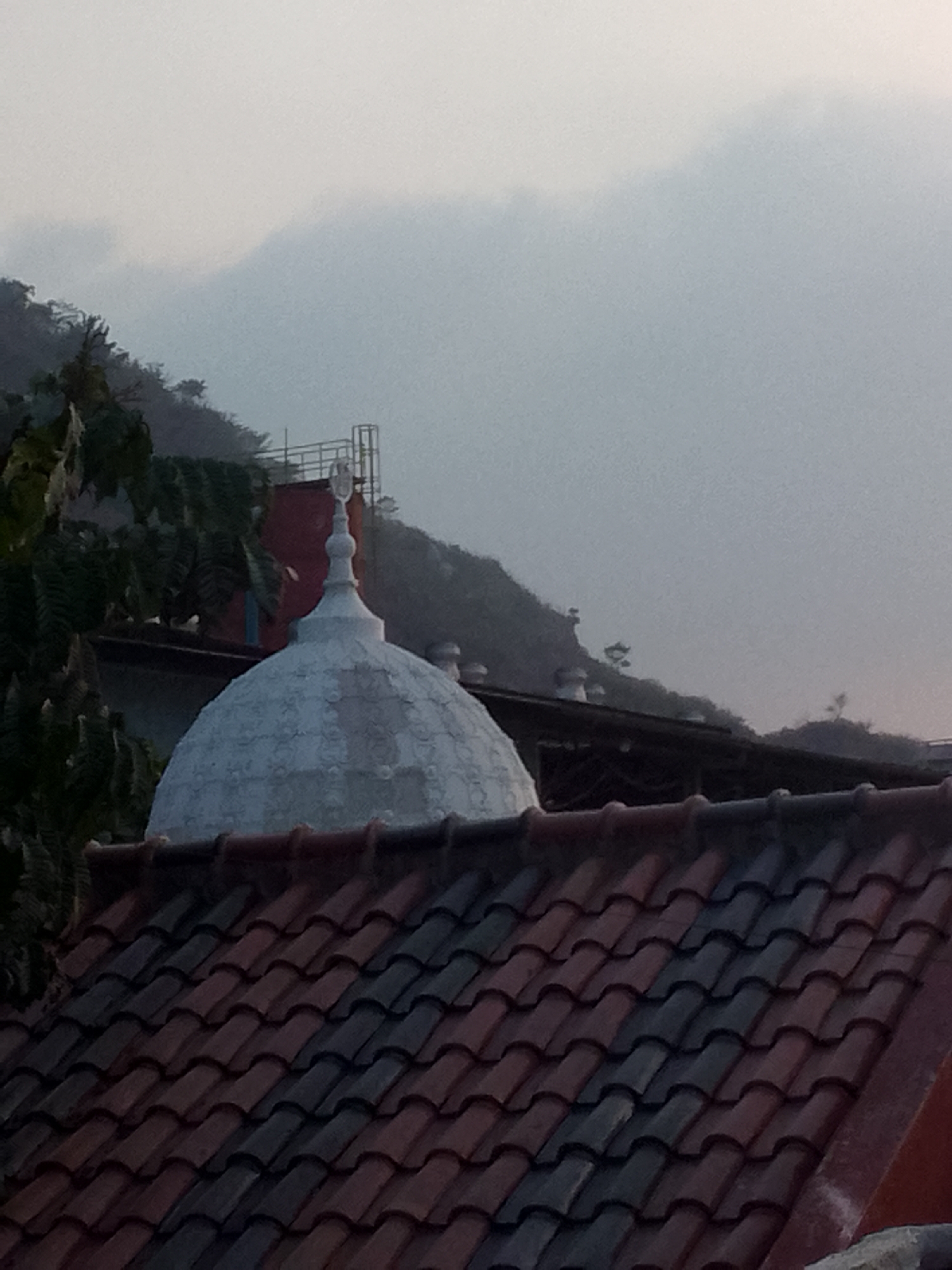 This image, taken in the vicinity of Gunungmasigit, Indonesia, depicts a hazy, overcast sky with subtle contours of a distant mountainous landscape. In the foreground, a tiled roof, composed of reddish-brown and dark gray tiles arranged in overlapping rows, dominates the lower portion of the frame. Perched above the roofline is the white dome of a structure, possibly a mosque, topped with a crescent moon finial. To the left of the dome, dark green foliage from a tree extends into the frame. Behind the dome, a red building is partially visible, and further in the background, a metallic structure resembling scaffolding or a communication tower can be seen against the misty hillside. The overall atmosphere is muted due to the low light and atmospheric haze.