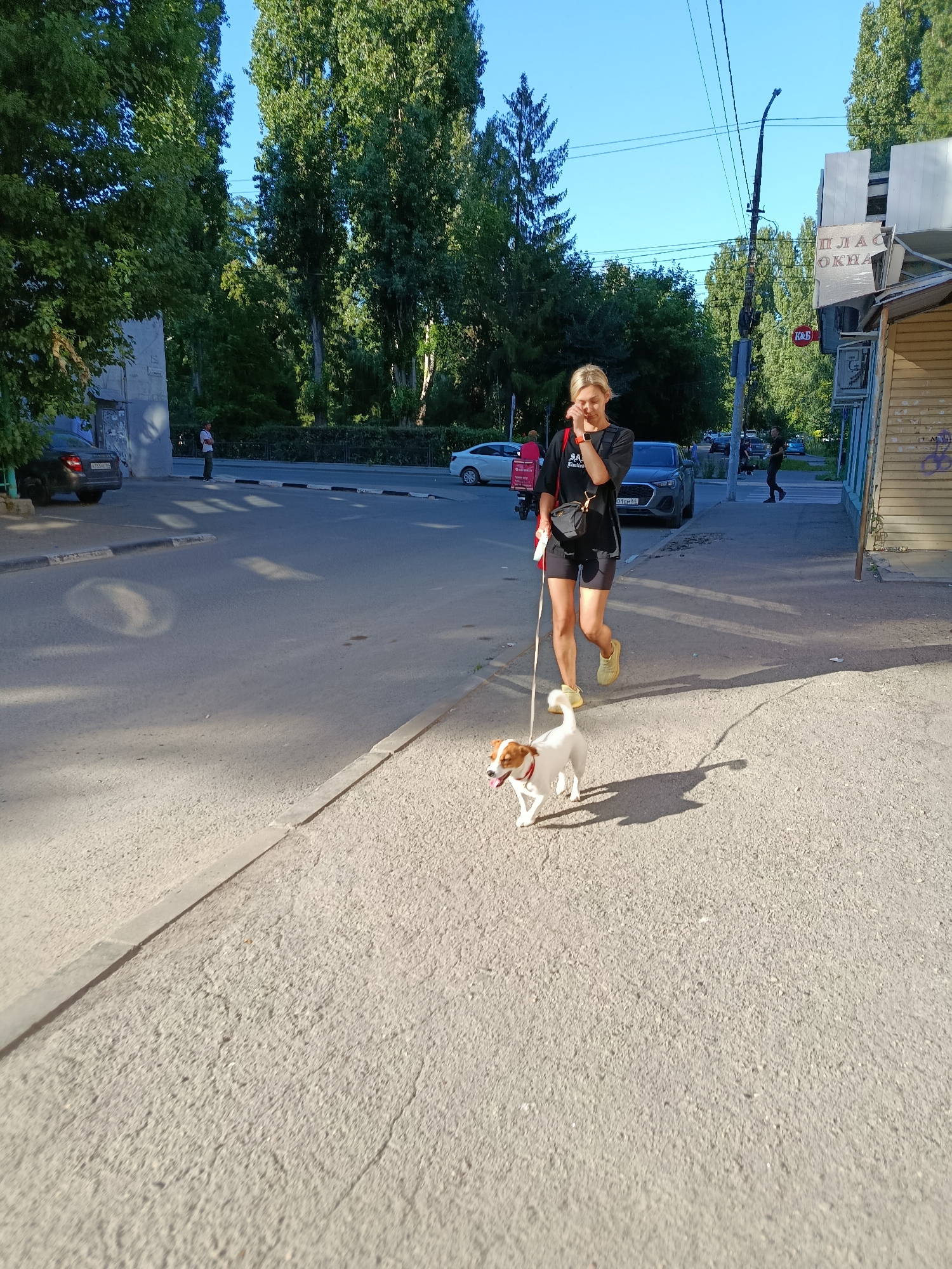 A blonde woman walks a small, white and tan dog on a leash. The woman wears a black t-shirt, black shorts, and yellow sneakers. She has a small black bag slung across her chest. The dog, a Jack Russell Terrier, is trotting happily beside her. They are walking on a paved sidewalk next to a street. There are cars driving on the street and trees lining the sidewalk. In the background, there is a building with a sign that reads "ПЛАСТИКОВЫЕ ОКНА" (Plastic windows). The sun is shining, suggesting it is daytime. The scene appears to be in a residential or commercial area.