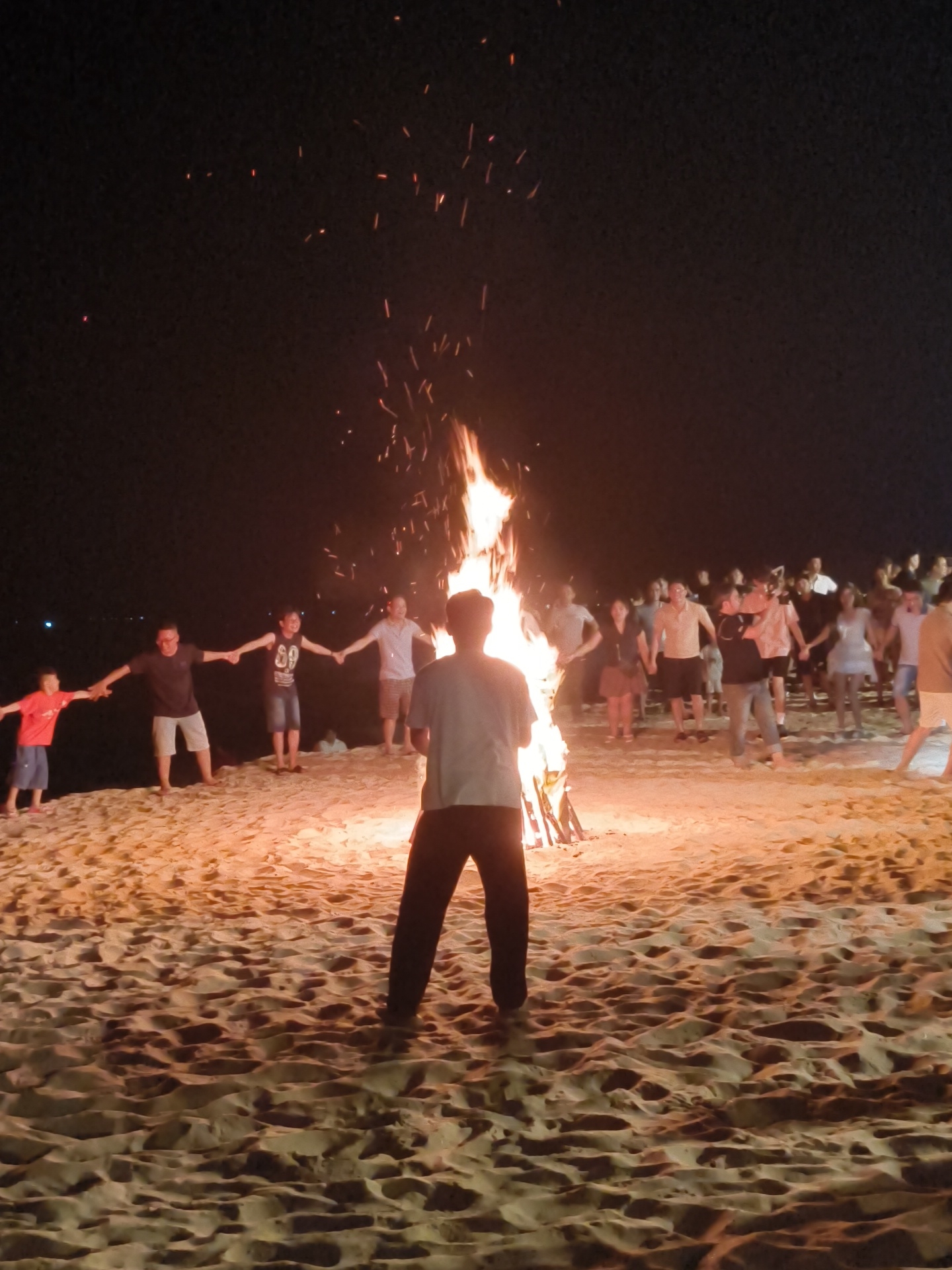 A group of people are gathered on a sandy beach at night, holding hands in a circle around a large bonfire. The fire casts a warm glow on their faces and the surrounding sand. Sparks from the fire ascend into the dark night sky. The people appear to be enjoying themselves, with some smiling and looking towards the fire. The scene suggests a celebration or a social gathering. The time of day is clearly night, as evidenced by the dark sky and the illuminated bonfire. The location appears to be a beach, with sand as the dominant ground surface. The overall mood is festive and communal.