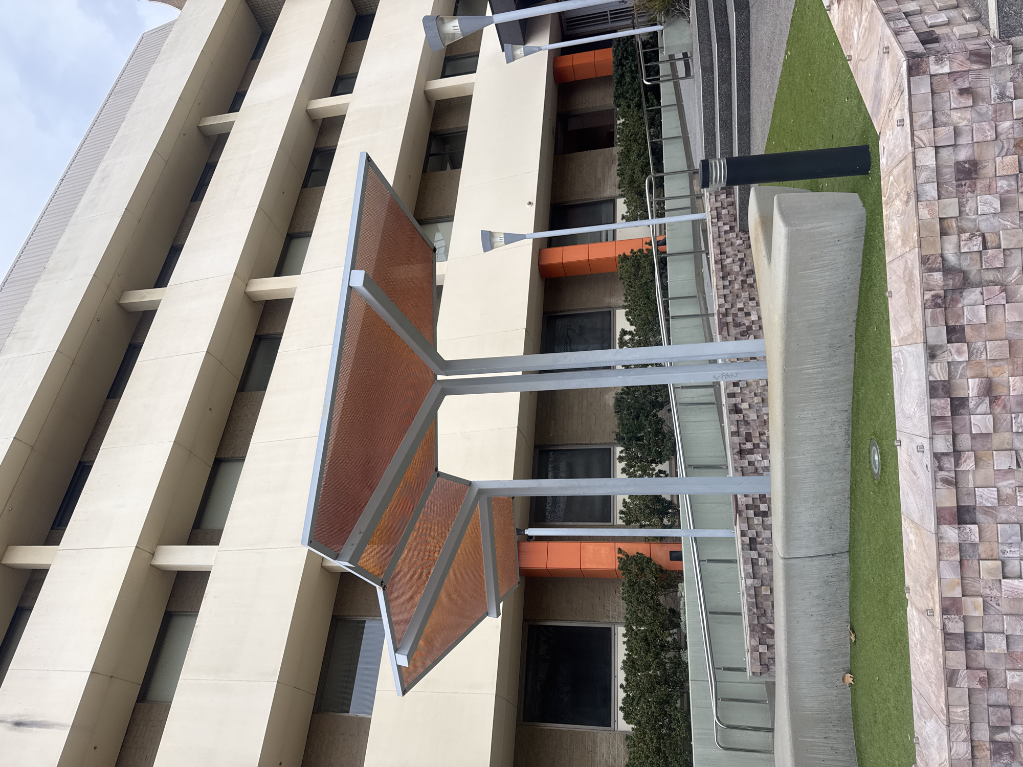 The image shows the exterior of a modern building with a light-colored facade, featuring multiple levels of balconies and windows. In the foreground, there is a contemporary outdoor seating area with a curved concrete bench, artificial grass, and decorative stone walling. A prominent feature is an orange-tinted canopy supported by metal poles, providing shade over the seating area. Several modern lampposts are also visible. The overall setting appears to be an urban environment, likely a public space or a corporate campus. The building's architecture and the landscaping suggest a well-maintained and recently developed area. The sky is overcast, indicating a cloudy day.