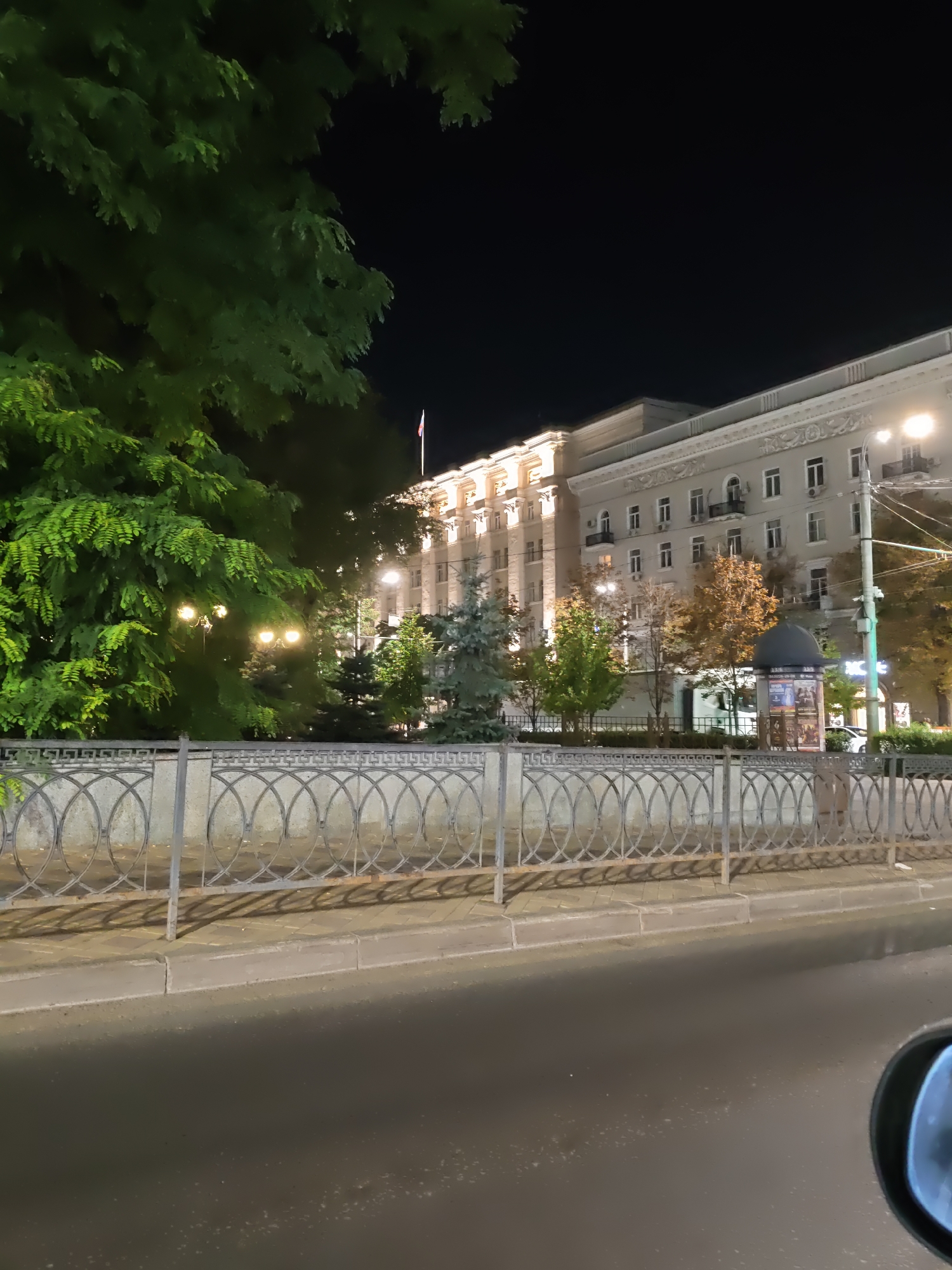 The image captures a nighttime view of a street scene in Rostov-on-Don, Russia. In the foreground, a wet asphalt road reflects the streetlights. A metal fence with an oval pattern separates the road from a sidewalk paved with light-colored rectangular tiles. Beyond the fence, several trees, some with lush green foliage and others with autumnal yellow and brown leaves, are visible.
Dominating the background is a large, multi-story building with a classical architectural style, illuminated by artificial lights. The building features a light-colored facade with numerous windows, balconies, and decorative cornices. A flag flies from the top of the building. Streetlights are positioned along the street and within the park-like area in front of the building. A dark, dome-shaped structure, possibly a kiosk or bus stop, is situated near the building. The overall atmosphere is one of a city at night, with artificial lighting creating a dramatic contrast with the dark sky.