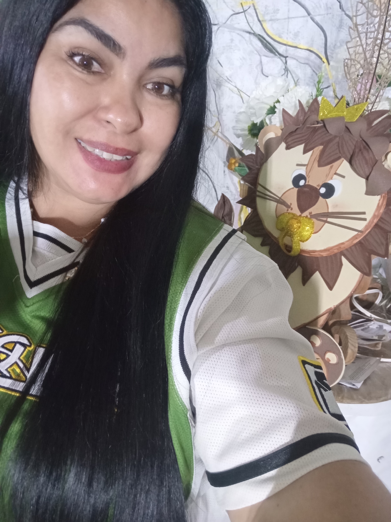 A woman is taking a selfie indoors with a decorative lion figure. The woman has dark hair and is wearing a green and white jersey-style shirt. The lion figure is made of layered cardstock or similar material, with a crown and a shiny pacifier-like object in its mouth. The background includes white floral decorations. The location is unknown and cannot be determined from the image.